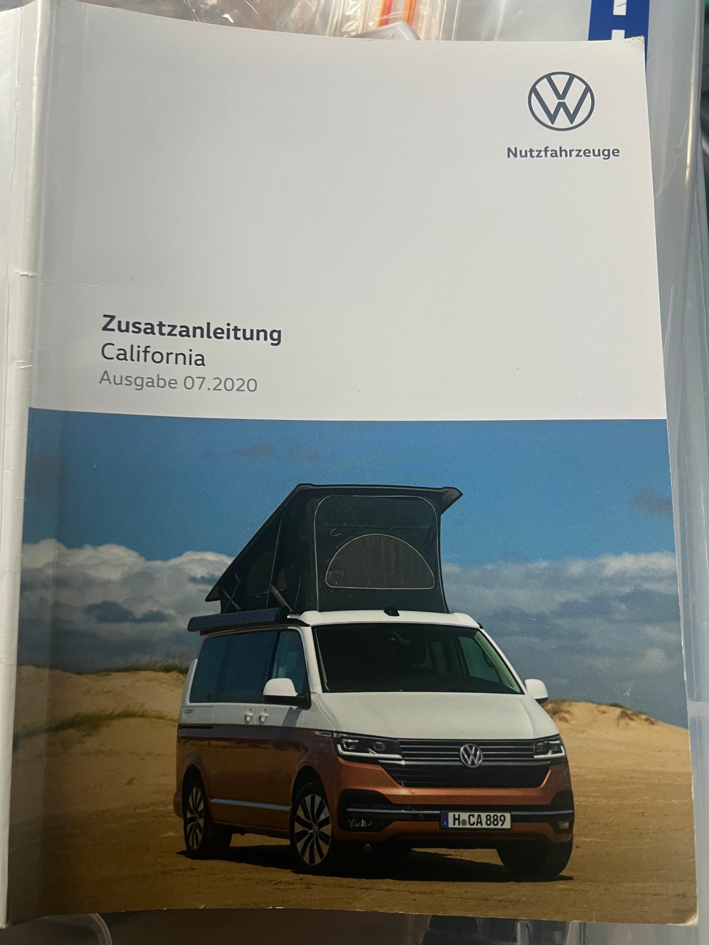 VW California camper van manual cover: white/brown van with open pop-up roof on sand.