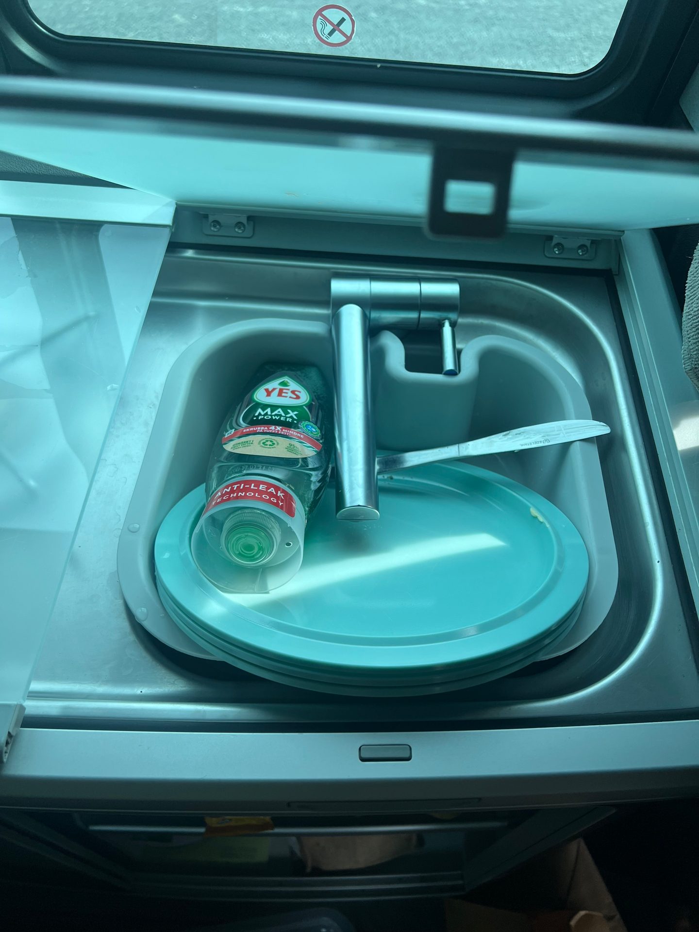 A small sink in a vehicle, containing dish soap, stacked turquoise plates, and a knife.