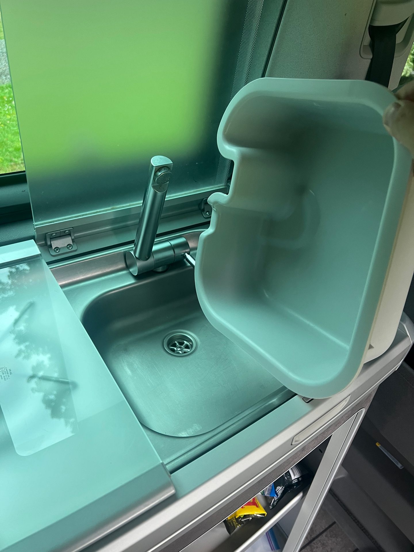A hand holds a light green plastic tub above a stainless steel sink and faucet in a camper van.