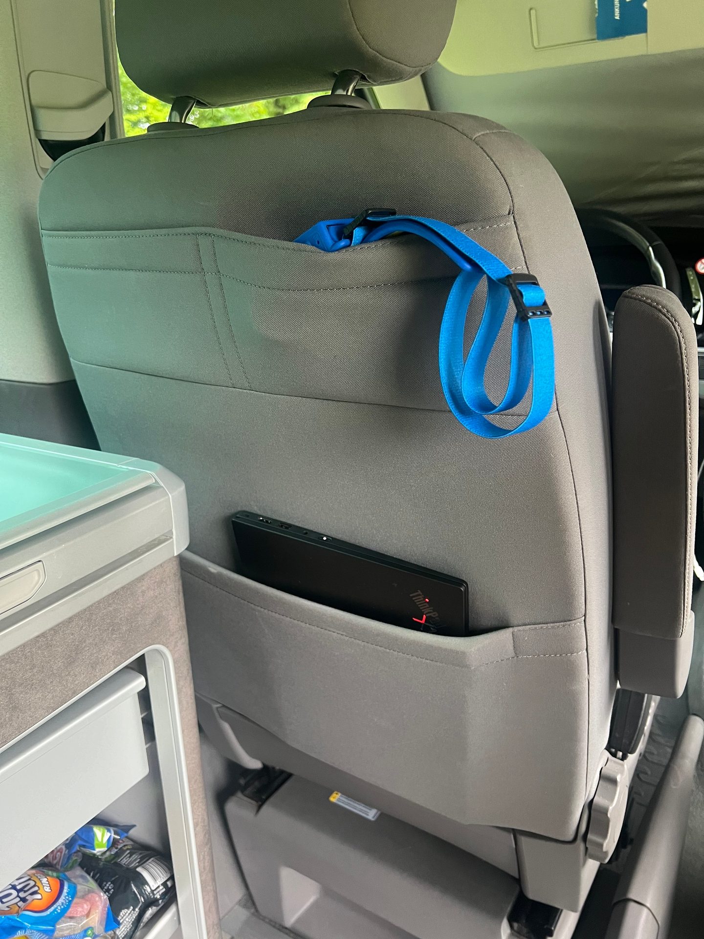 Back of a car seat with a laptop in a pocket, a blue strap, and a cooler next to it.