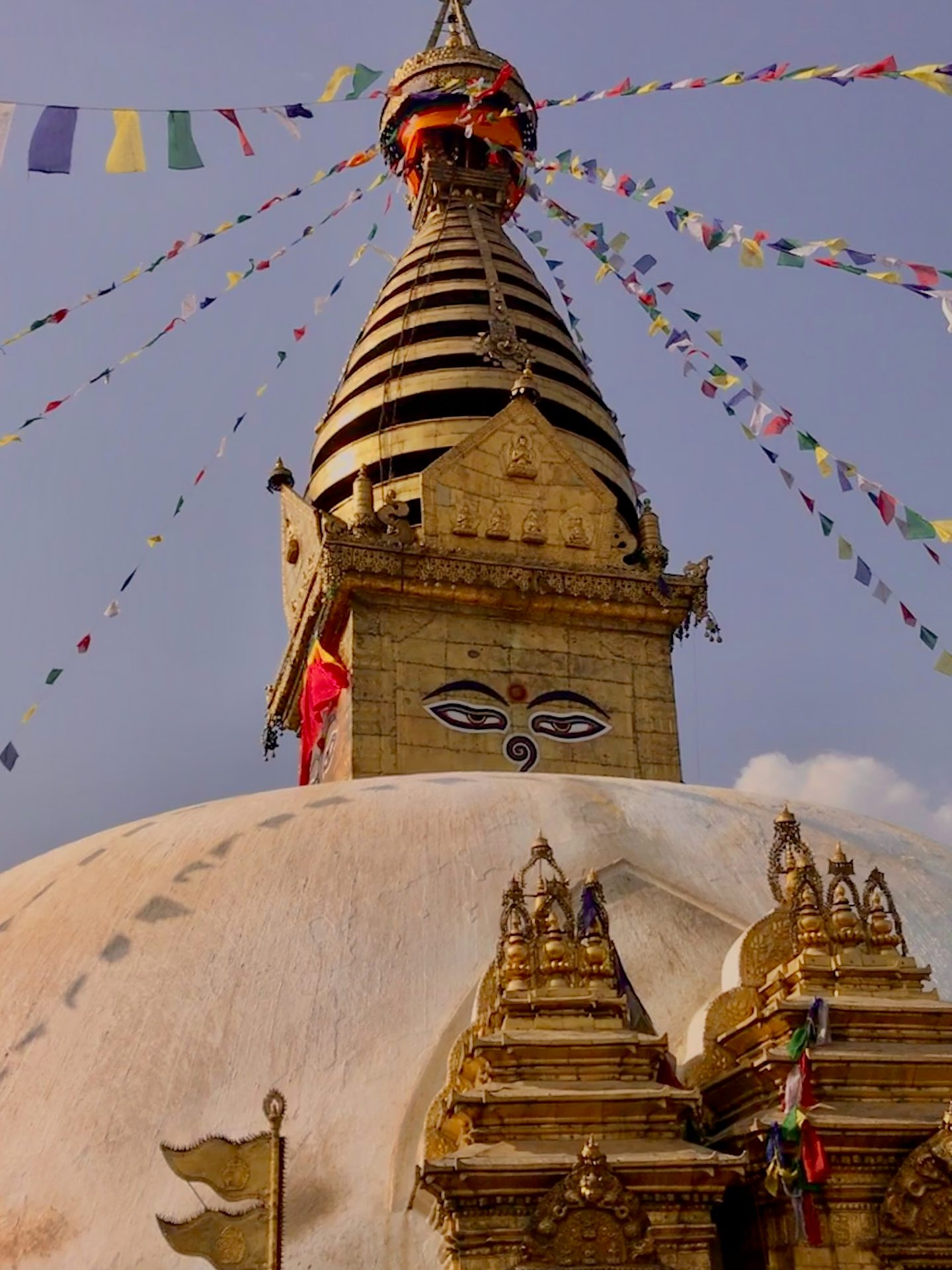 Golden stupa with painted eyes, white dome, and colorful prayer flags against a blue sky.