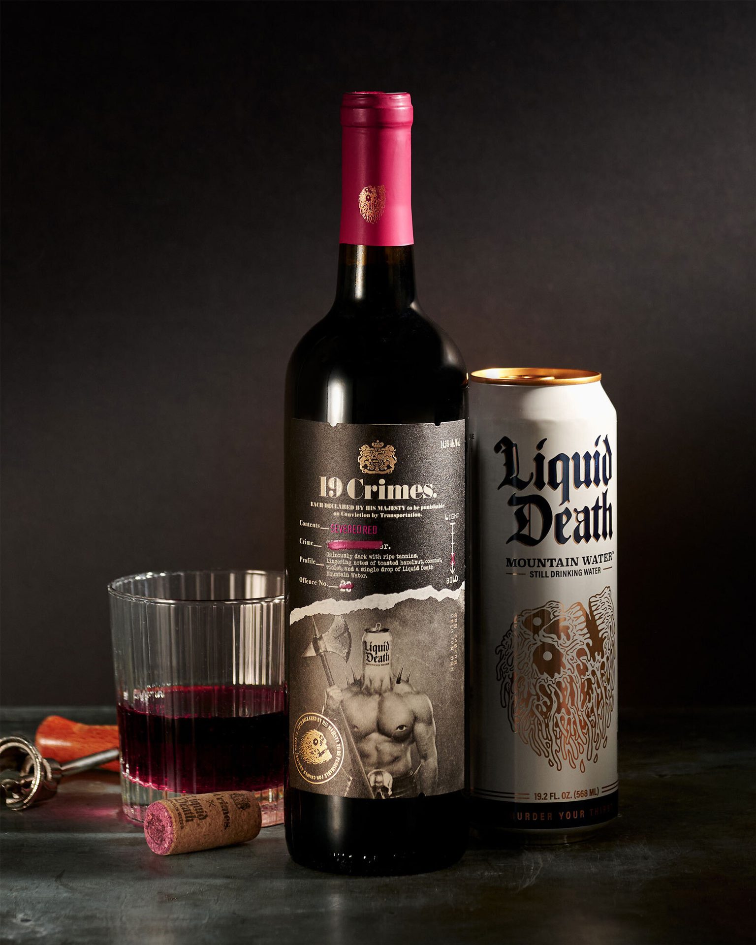 19 Crimes wine bottle, Liquid Death can, wine glass with red wine, and corkscrew on dark surface.