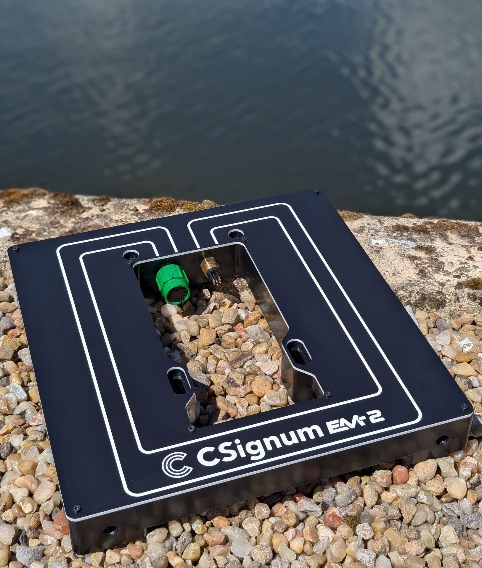 Black CSignum EM-2 device with green and brass connectors, resting on pebbles by water.