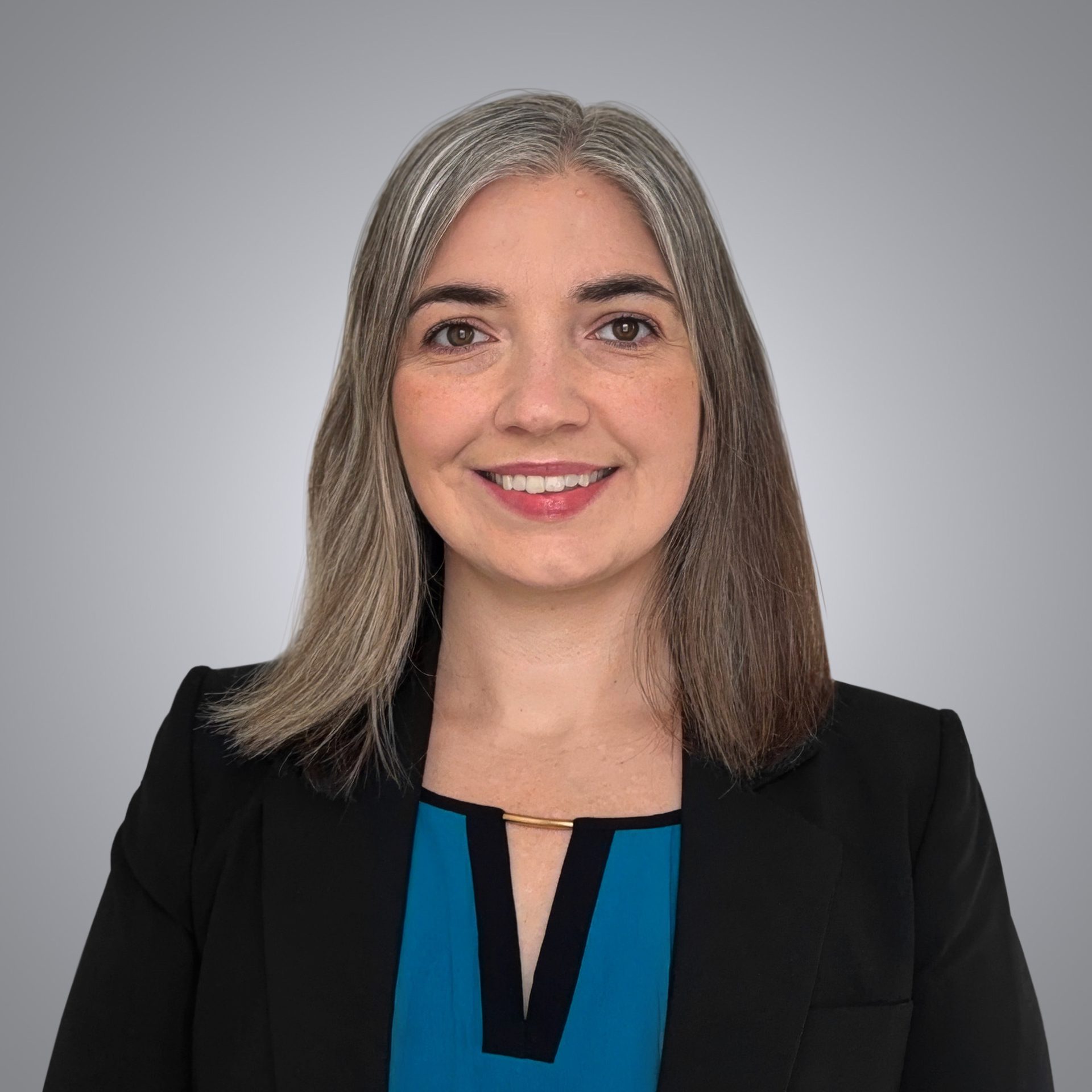 A smiling woman with long grey and brown hair wears a black blazer and blue top, looking directly at the camera.