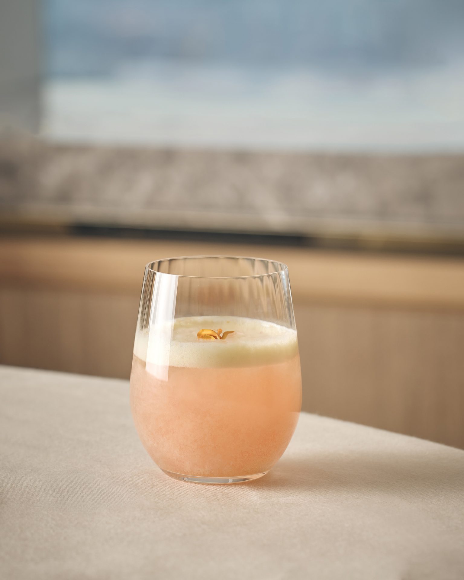 A short glass with a pink beverage, white foam, and an orange garnish on a light tablecloth.