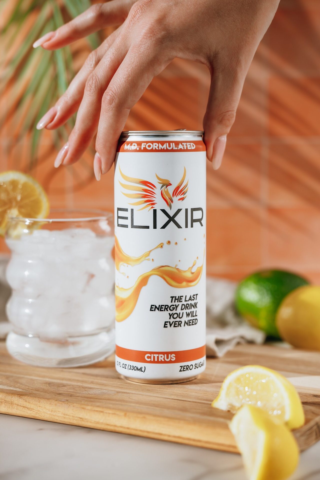 Hand holding Elixir Citrus energy drink can, with a glass of ice water, lemons, and lime.