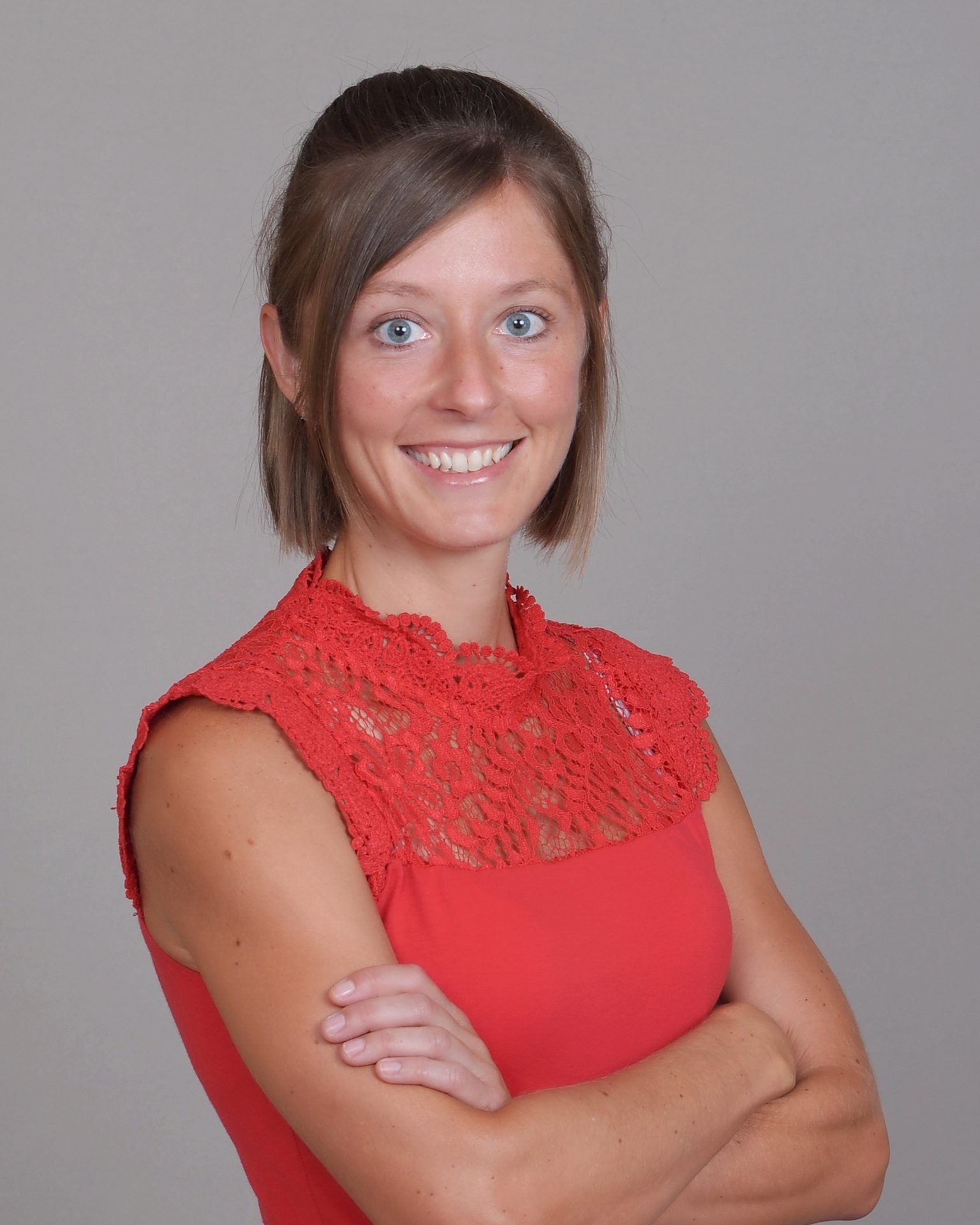Young woman with short brown hair, blue eyes, and a red lace-detail top, smiling with arms crossed.