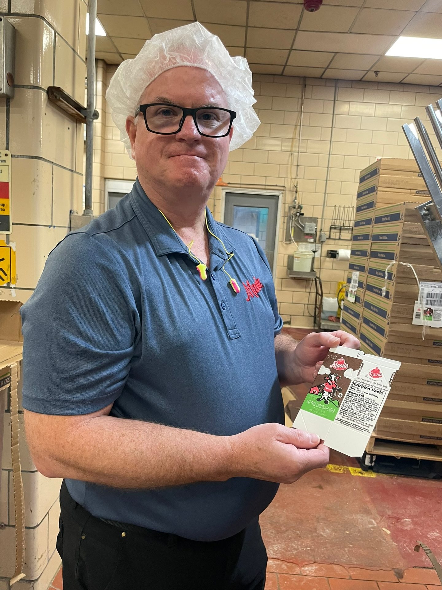 A man in a hairnet and glasses holds a chocolate milk carton in a factory.