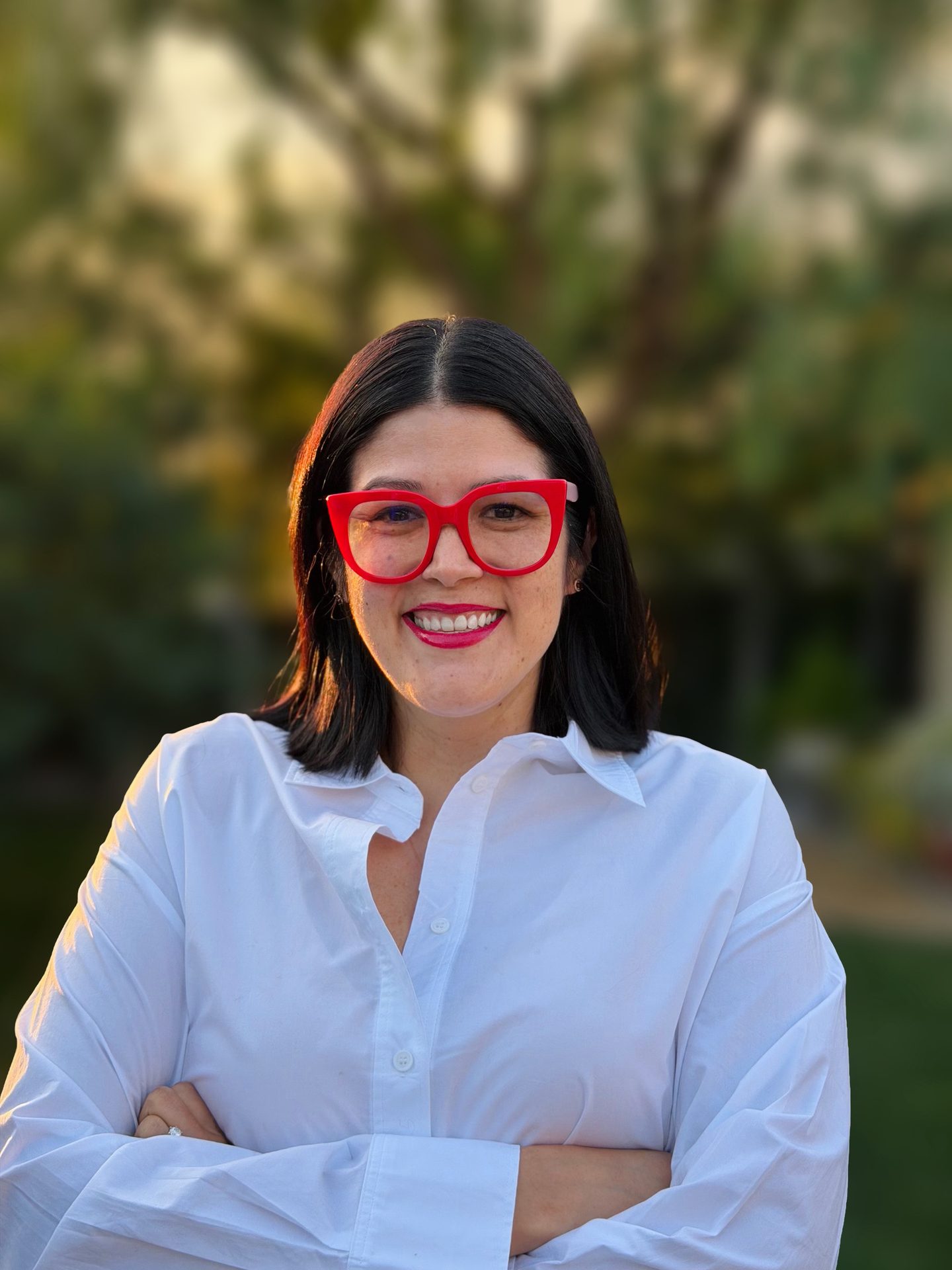 A smiling woman wearing red glasses and a white collared shirt, outdoors with a blurred green background.