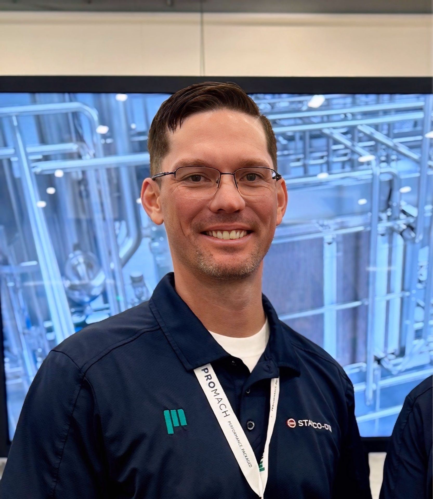 Smiling man in blue polo with logos and Promach lanyard, industrial background.