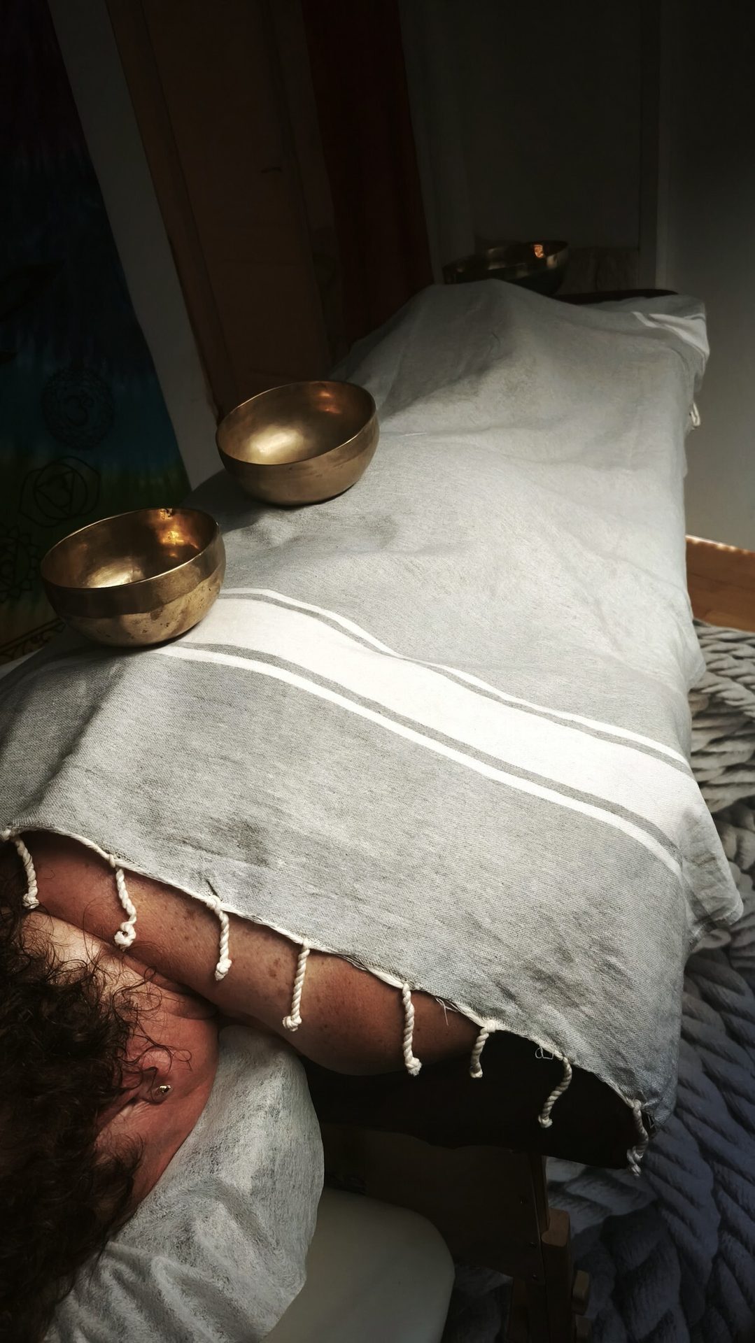 Person covered by a striped blanket, two brass singing bowls on their back.