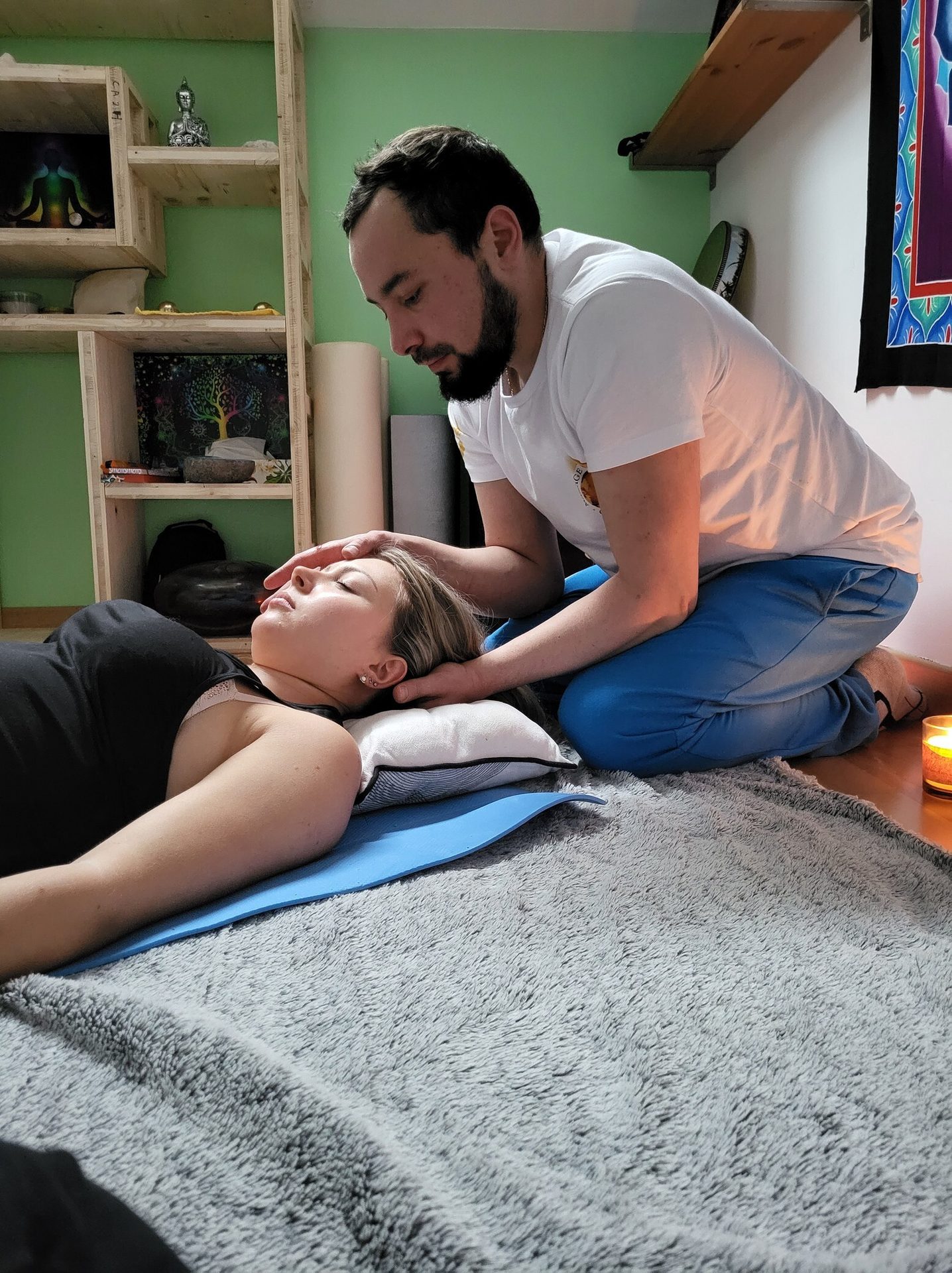 A man gives head and neck therapy to a woman lying on a mat in a calm, spiritual setting.