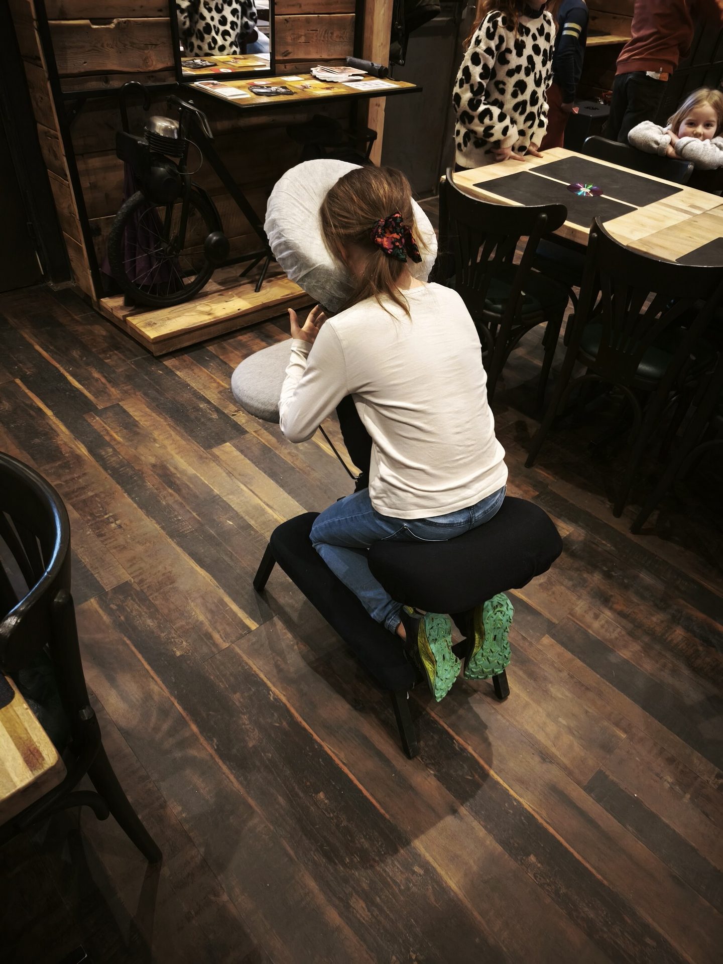 Person from behind in a portable massage chair, wearing jeans, in an indoor wooden space.