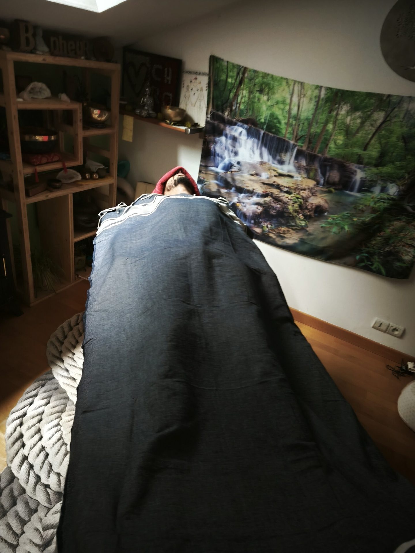 Person under dark blanket with red hood on rope cushion, wooden shelf and waterfall tapestry in background.
