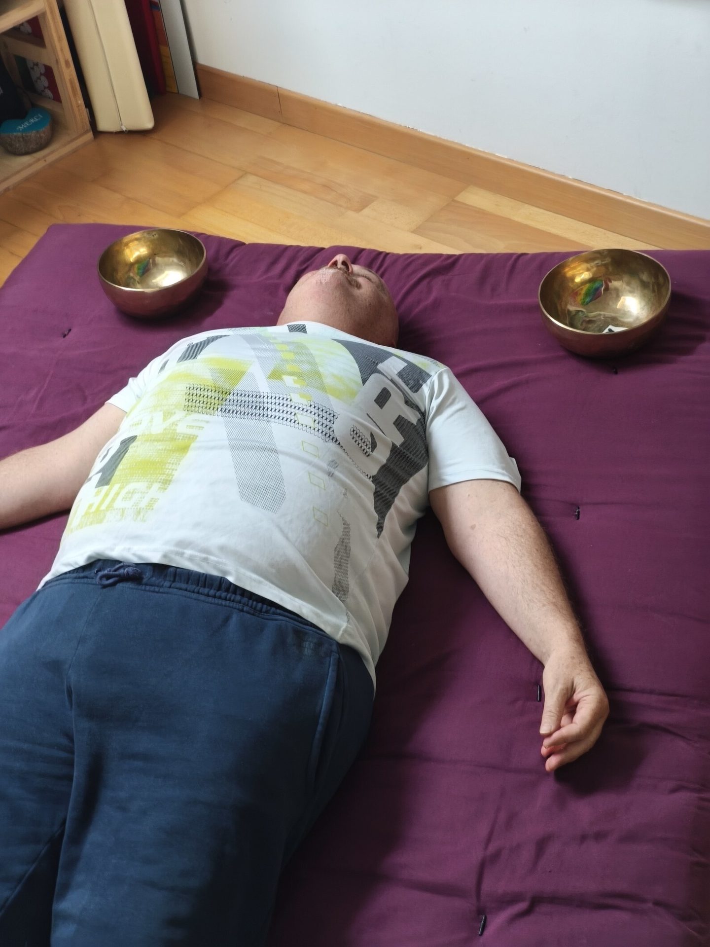 A man lies on a purple mat with two singing bowls near his head.