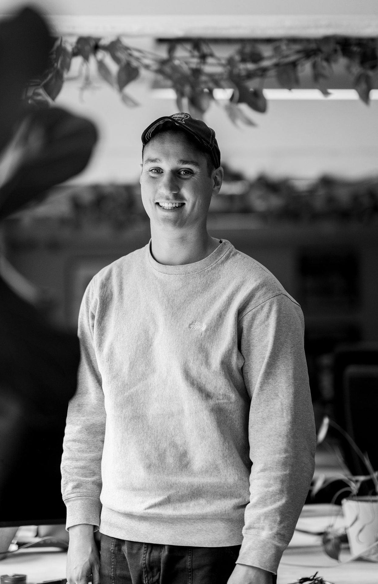 Black and white portrait of a smiling man wearing a cap and sweatshirt.