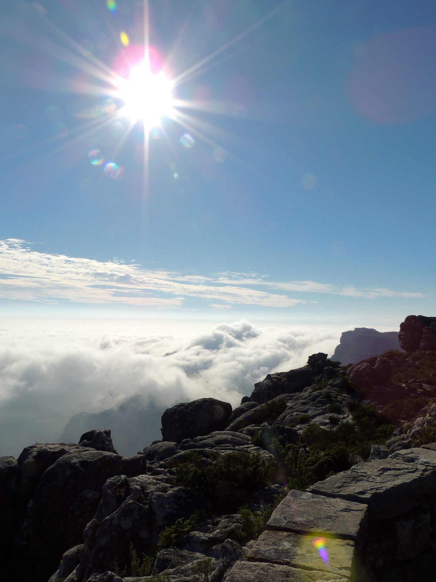 Bright sun over a sea of clouds, with rocky mountain peaks in the foreground and distant mountains.