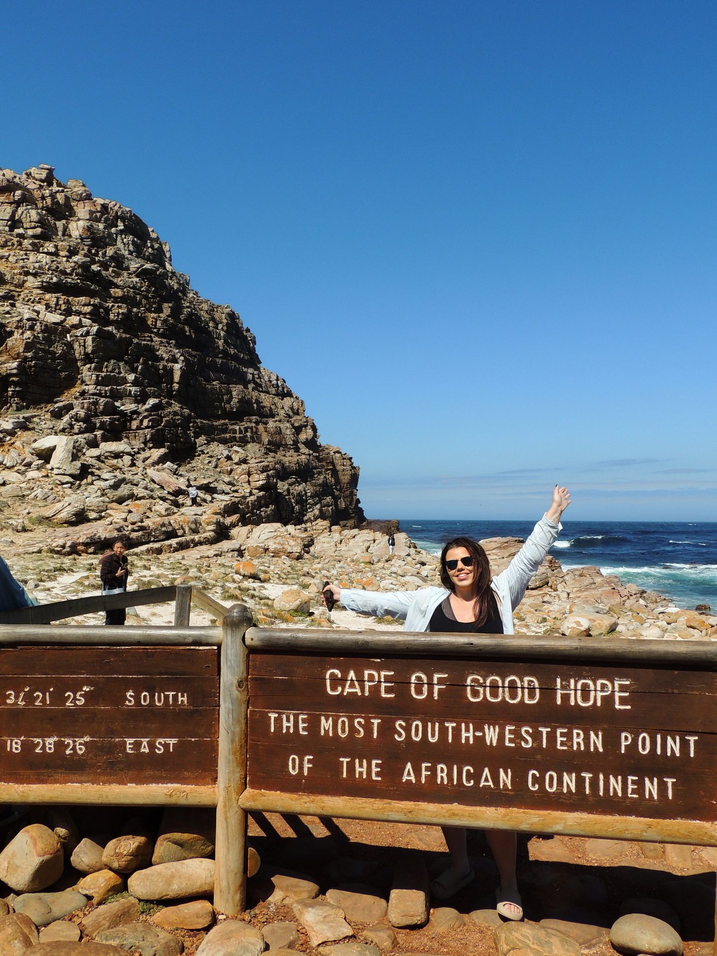 Woman posing by the Cape of Good Hope sign, with rocky cliffs and ocean under a blue sky.