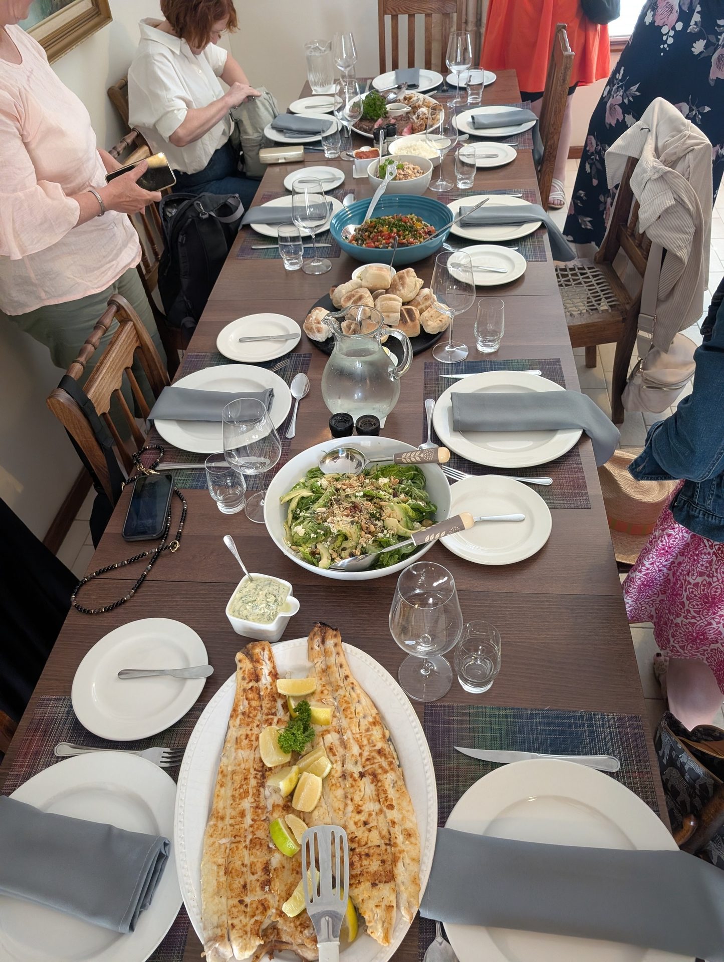 A dinner table laden with grilled fish, various salads, bread, and drinks, with people dining.