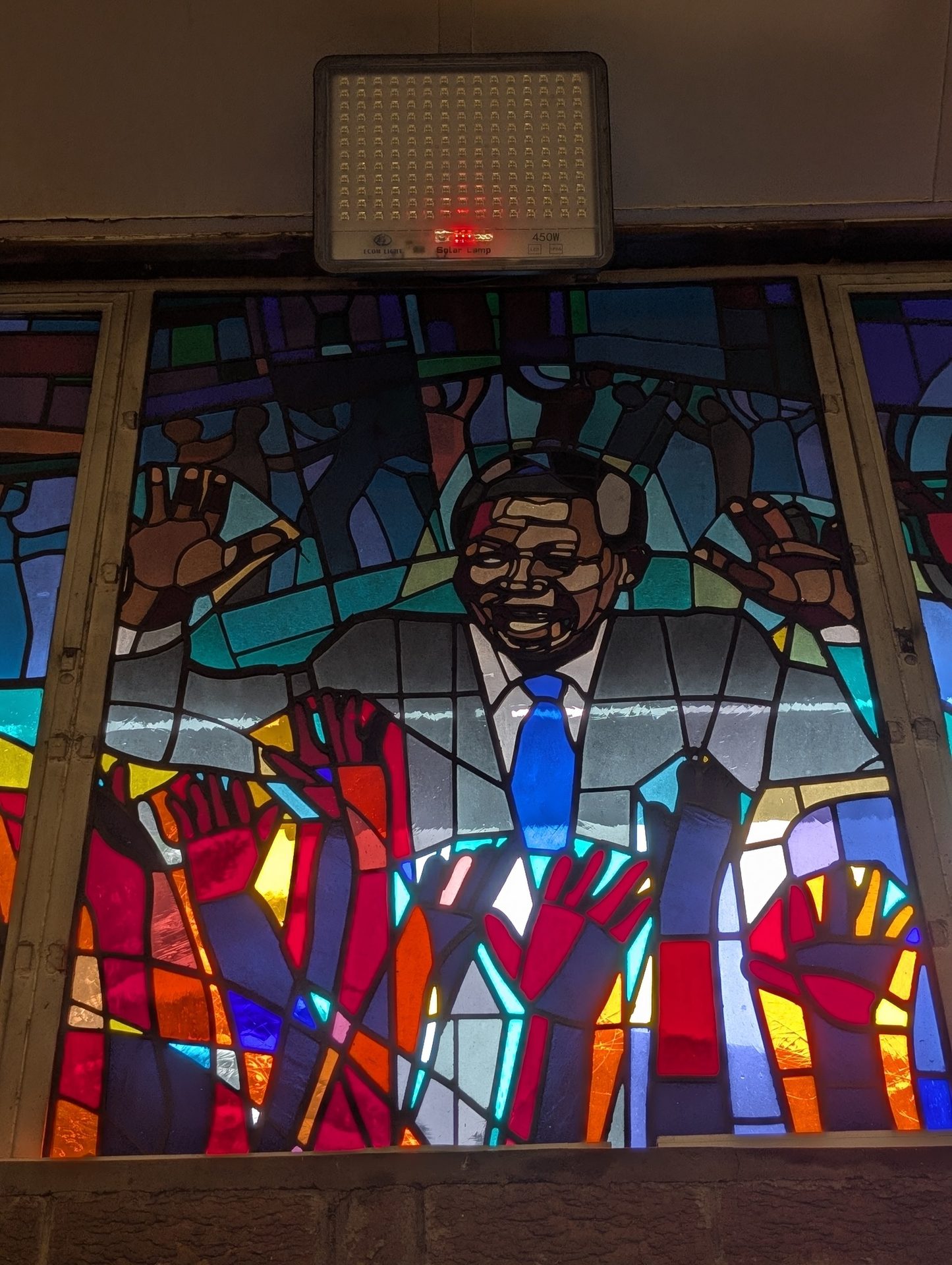 Stained glass window: smiling man, possibly Nelson Mandela, in suit and tie, with raised hands.
