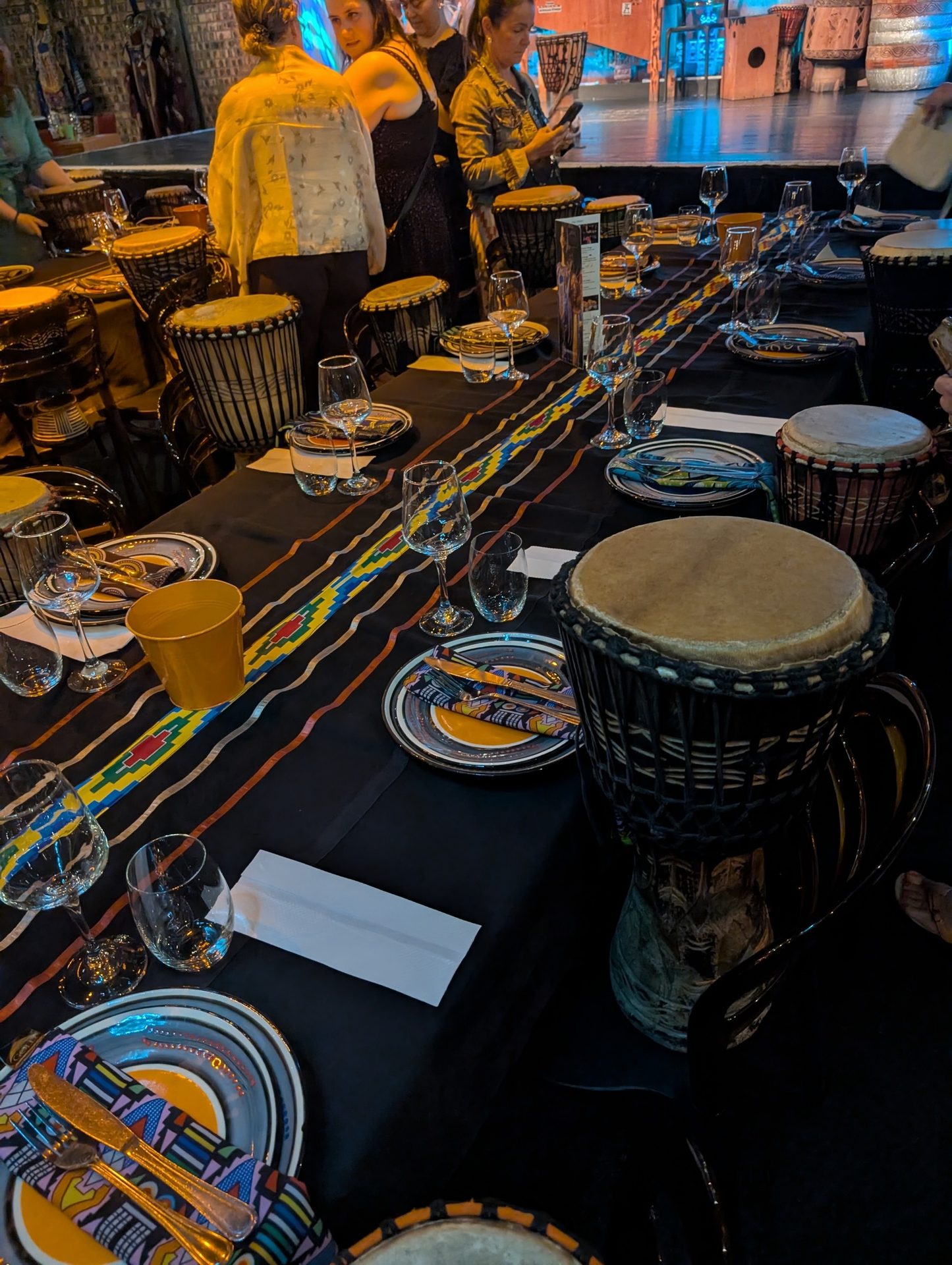 A long table with a colorful runner, set with plates, glasses, and numerous djembe drums. People gather around.