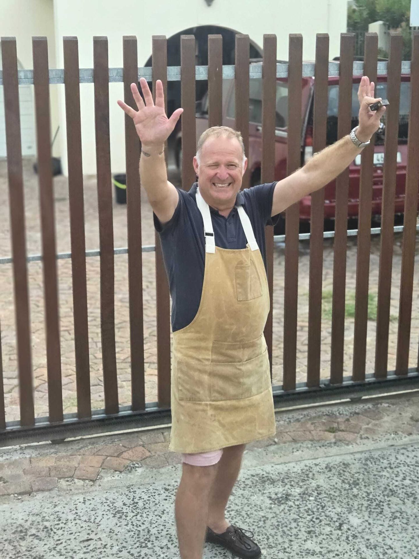 A smiling man in a brown apron waves cheerfully, standing in front of a wooden gate, holding keys.