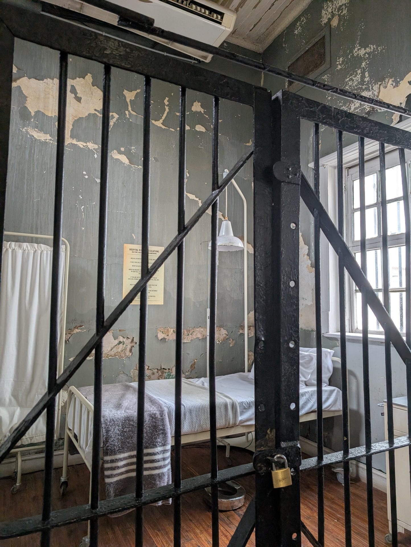 A dilapidated prison infirmary cell with a bed, medical stand, and peeling walls, viewed through iron bars.