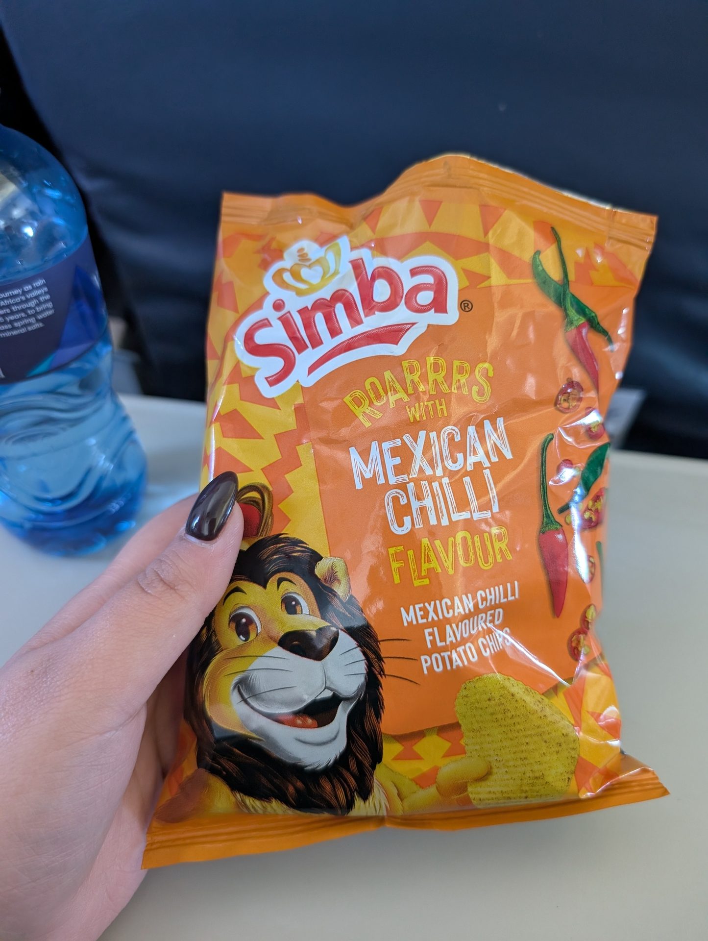A hand holds a vibrant orange bag of Simba Roarrrs Mexican Chilli Flavour potato chips, with a water bottle nearby.
