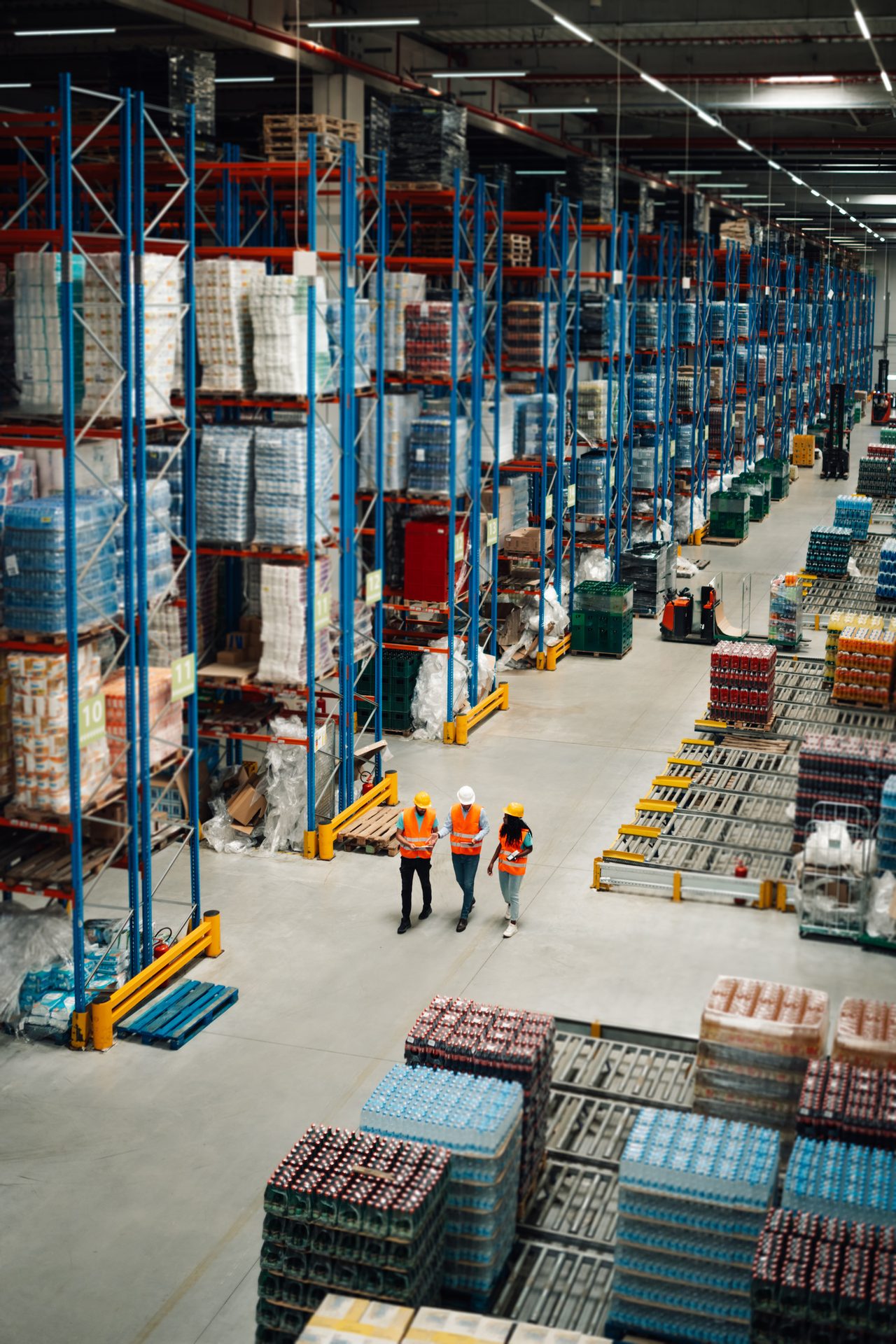 Three workers in safety vests and hard hats walk through a vast, organized warehouse with tall shelves.