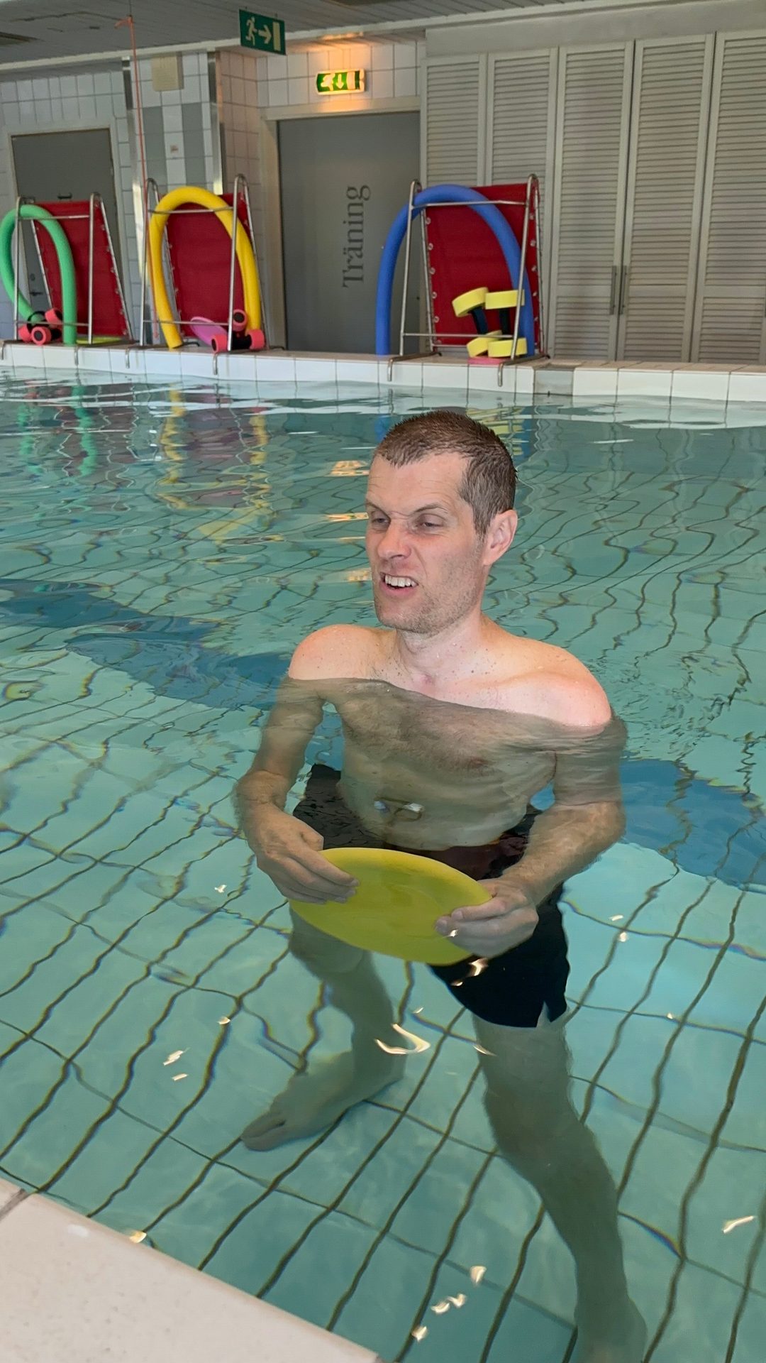 A man with short hair and a beard, wearing black swim shorts, stands in a swimming pool holding a yellow frisbee.