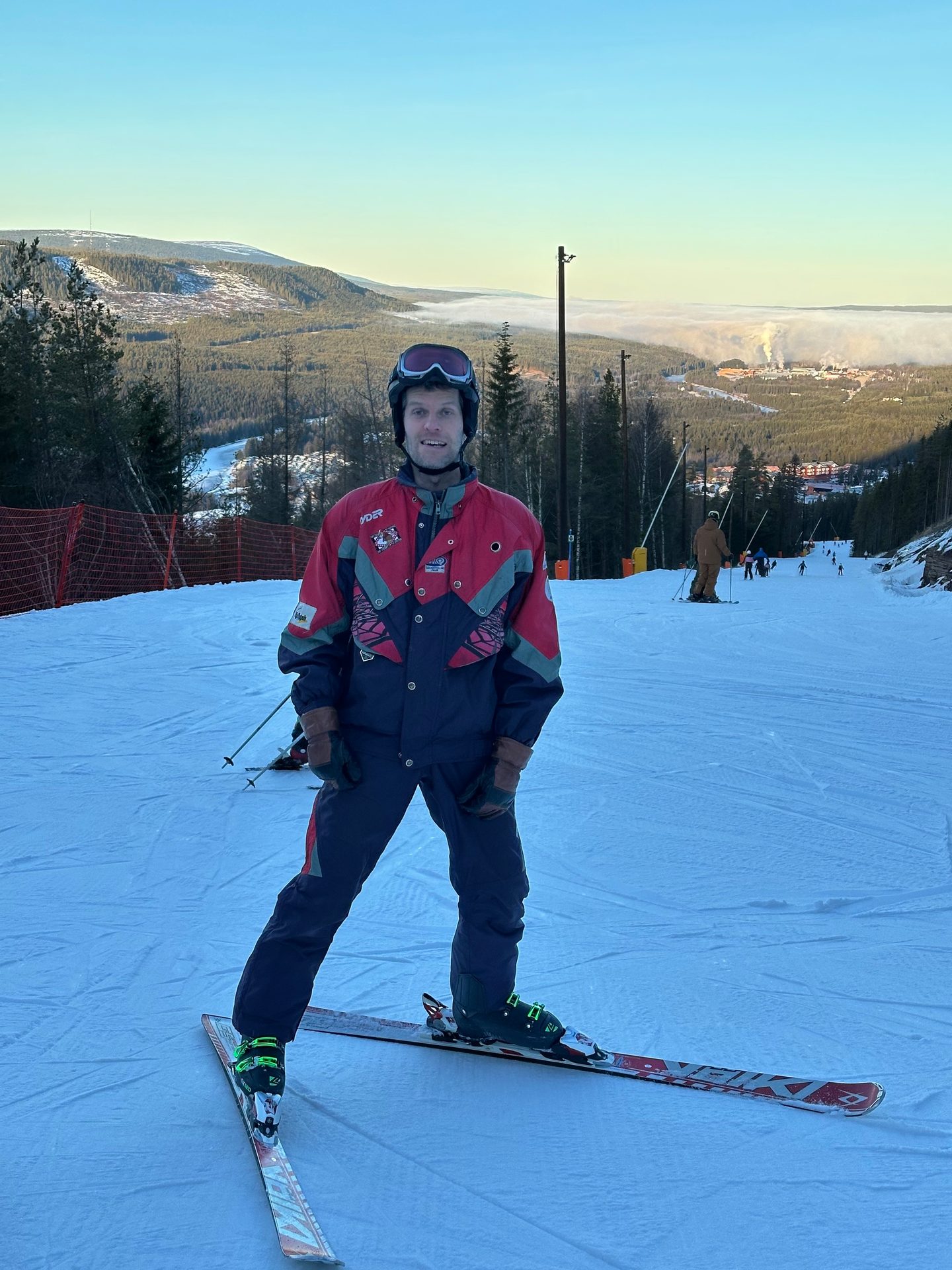 A man in a retro red and blue ski suit stands on skis on a snowy mountain slope with a foggy valley behind him.