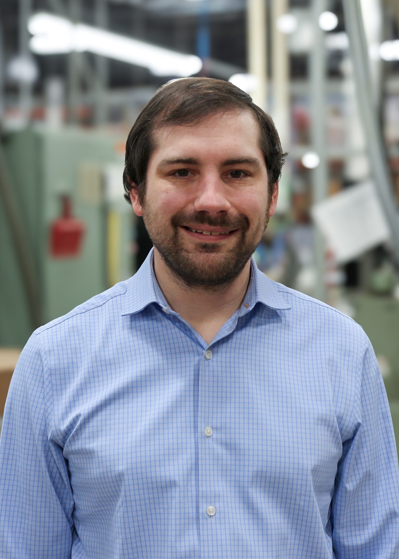 A man with a beard and dark hair smiles at the camera, wearing a blue checkered shirt in an industrial setting.