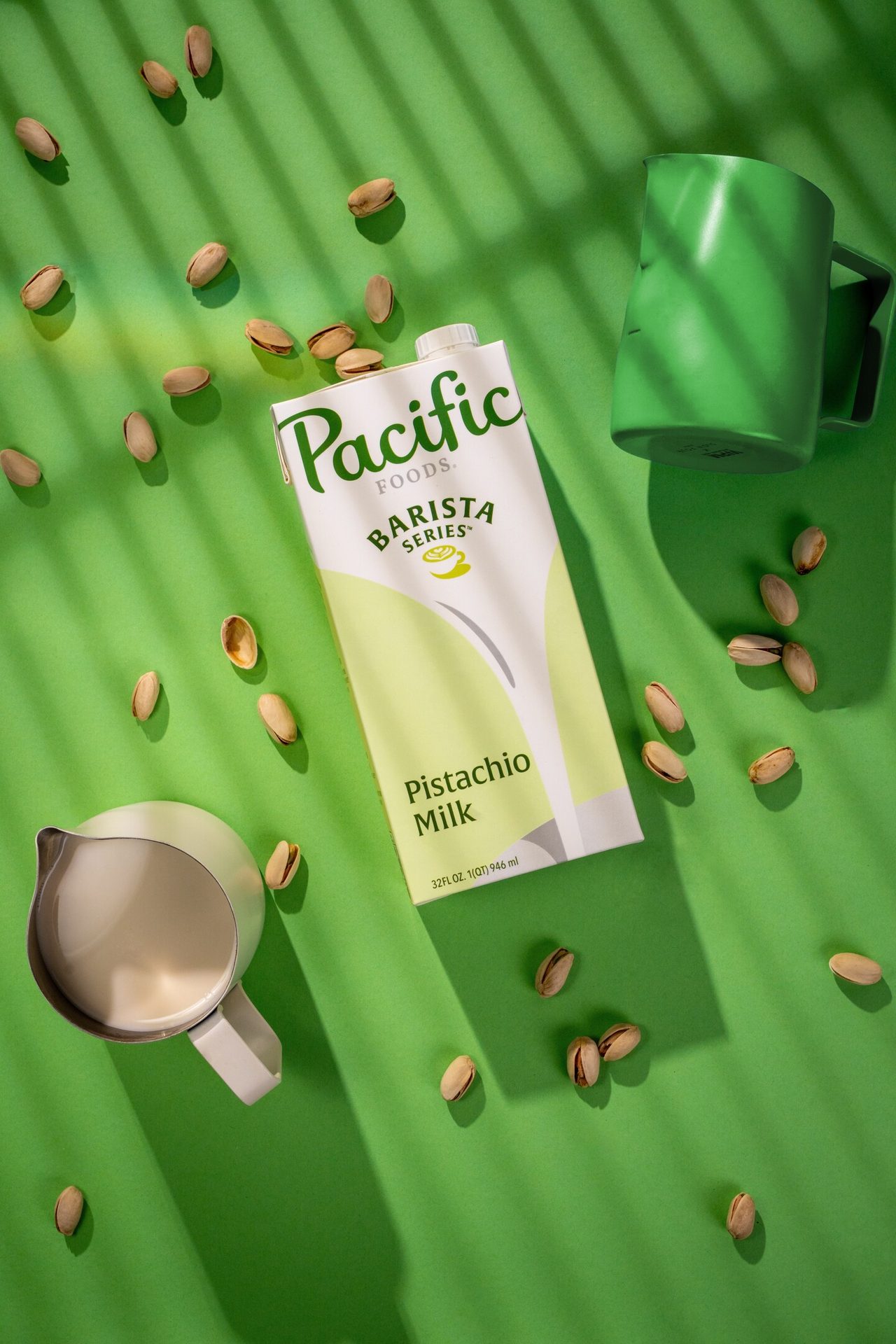 Pacific Foods Barista Series Pistachio Milk carton, pistachios, and two jugs on a green background with shadows.