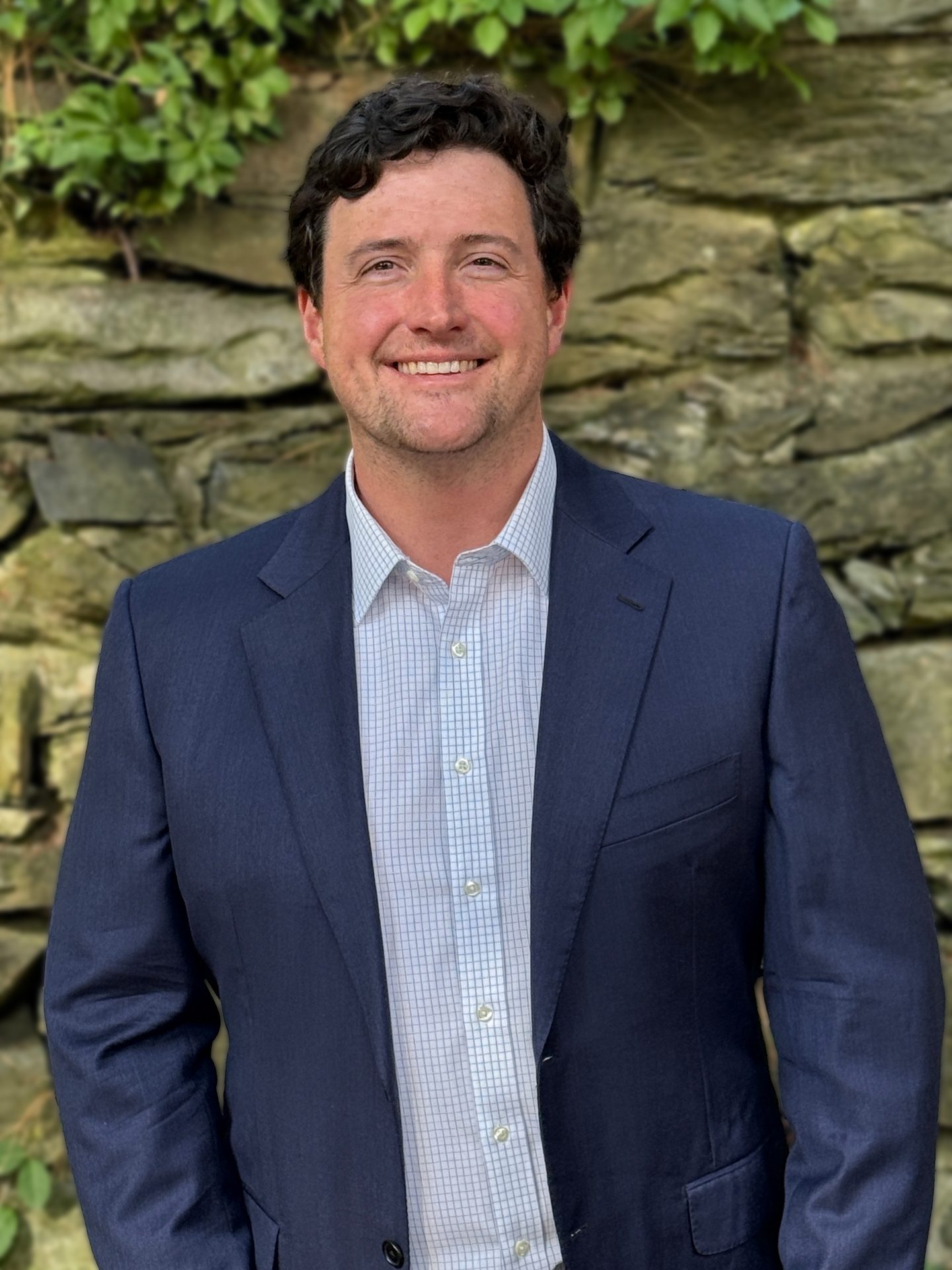 Smiling man in blue suit jacket and plaid shirt, standing against a stone wall with greenery.