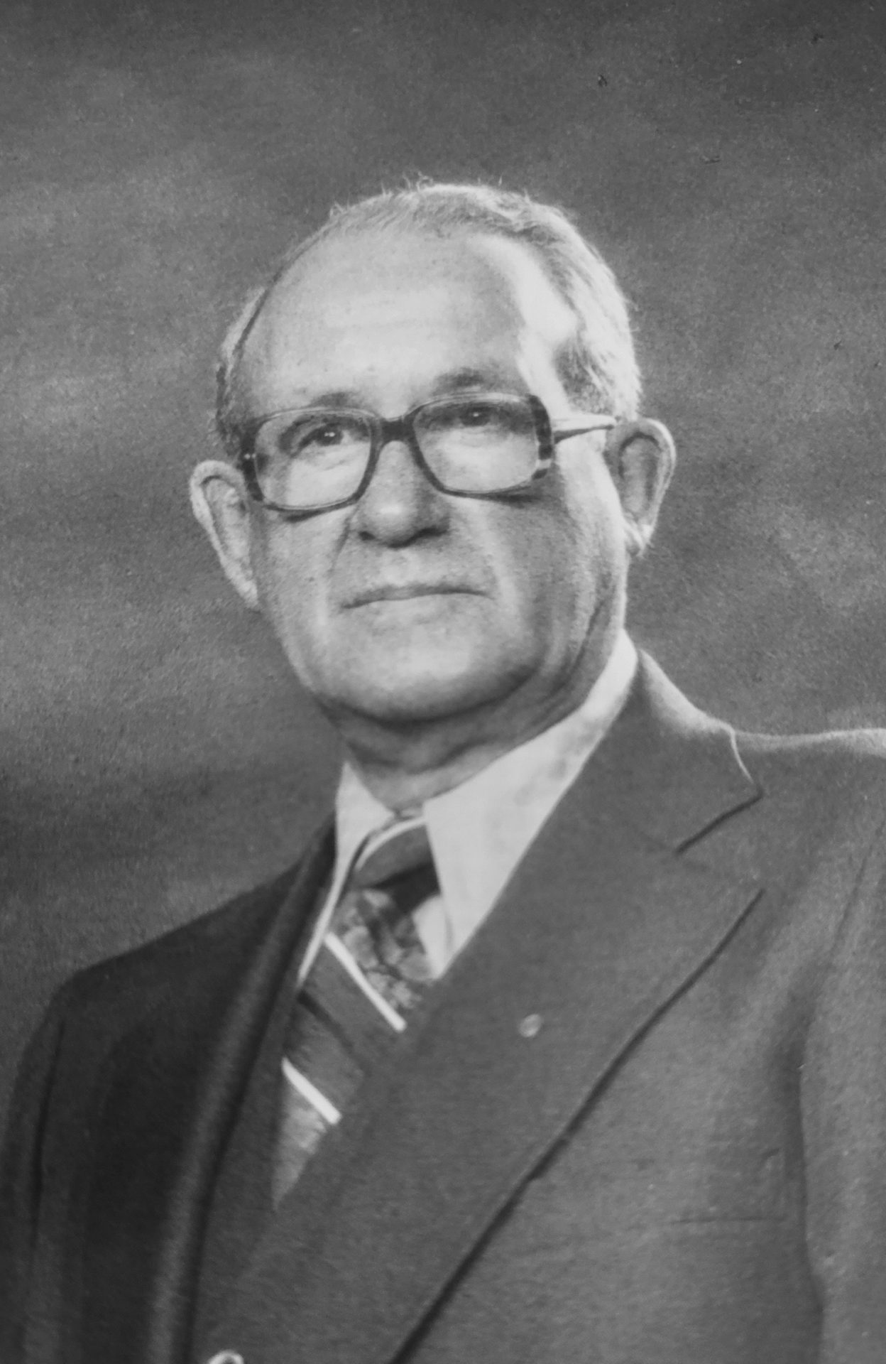 Black and white portrait of an older man with glasses, wearing a suit and tie.
