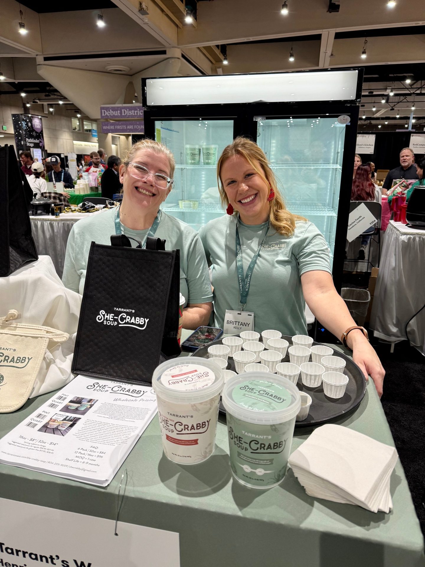 Two smiling women stand behind a table displaying "She-Crabby Soup" products at a trade show.