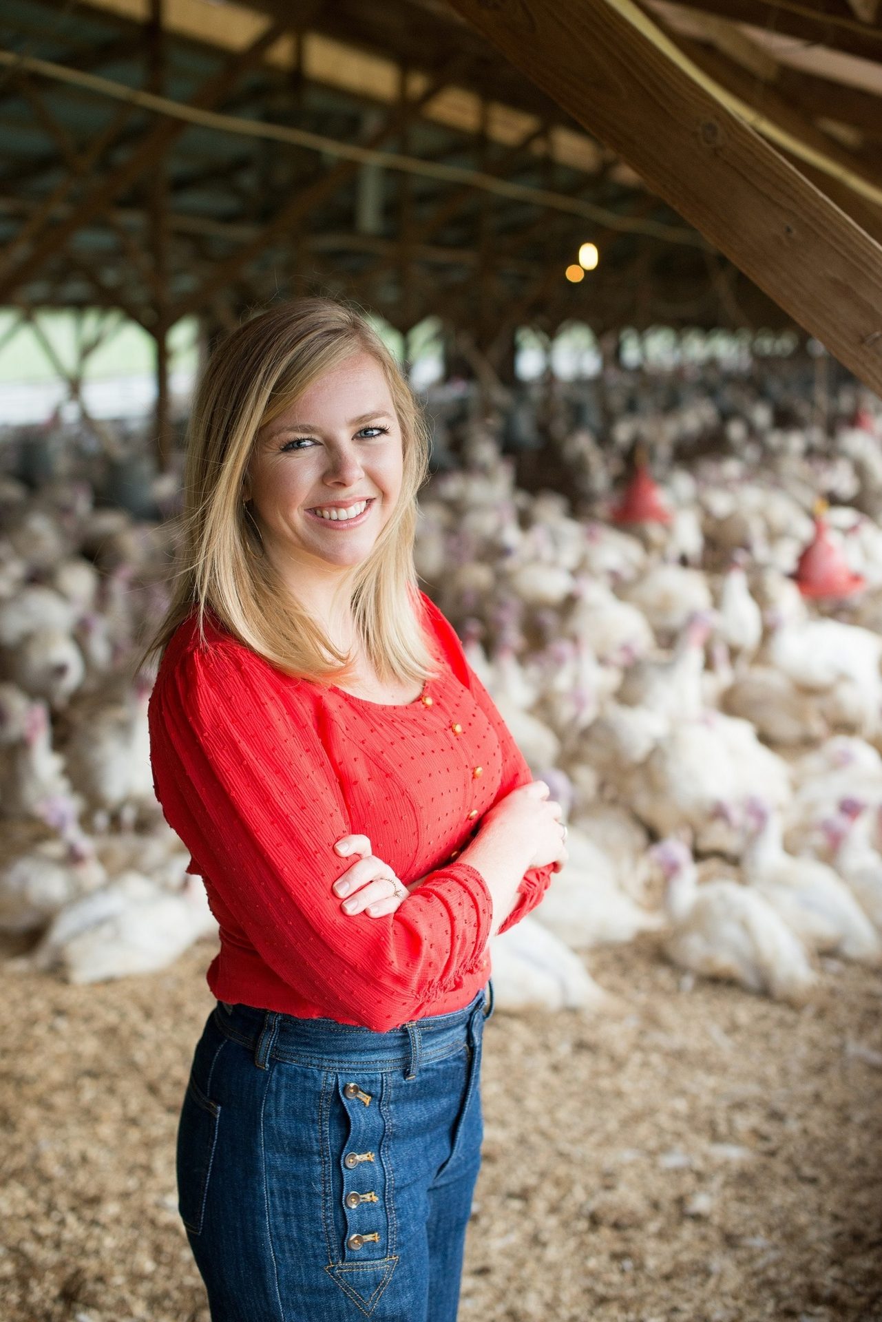 Smiling young woman in red shirt and jeans, arms crossed, in a barn full of turkeys.