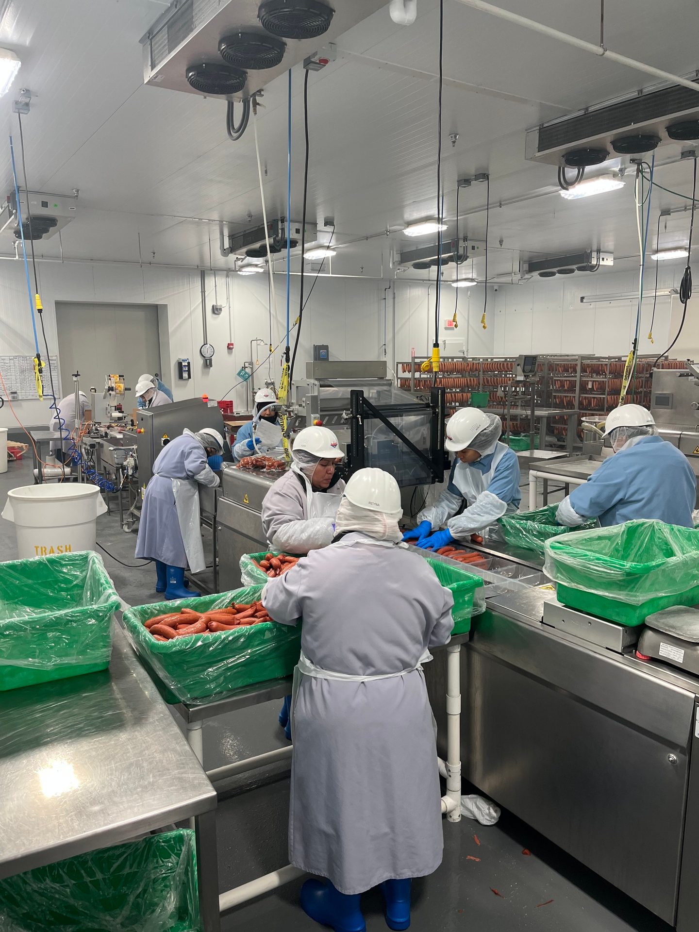 Workers in protective gear sort sausages in a clean food processing facility with machinery.