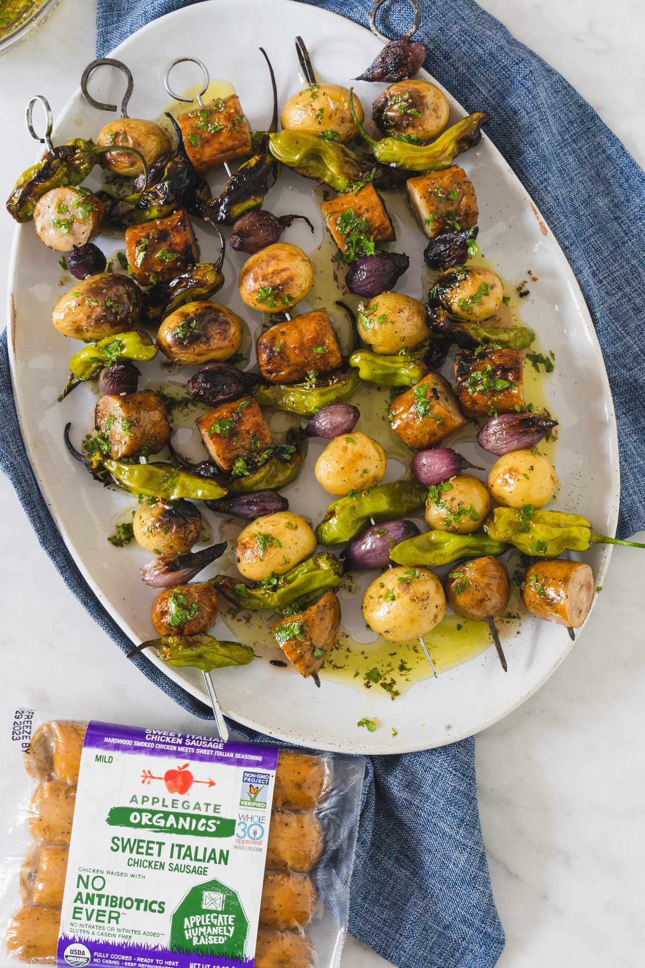 Grilled chicken sausage, potato, pepper, and onion skewers on a platter, with a sausage package.