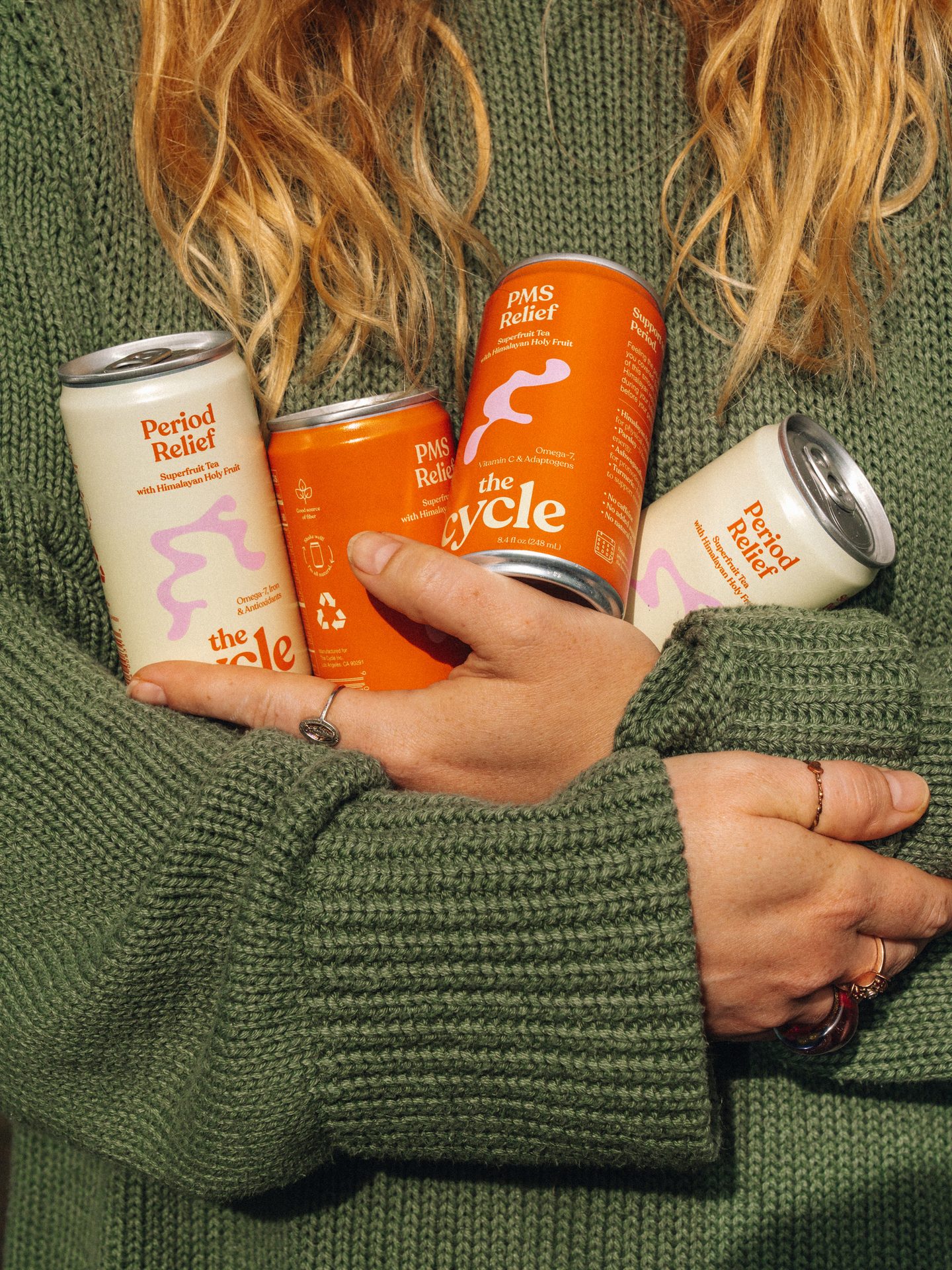 Person in green sweater holding four cans of Period Relief and PMS Relief drinks.