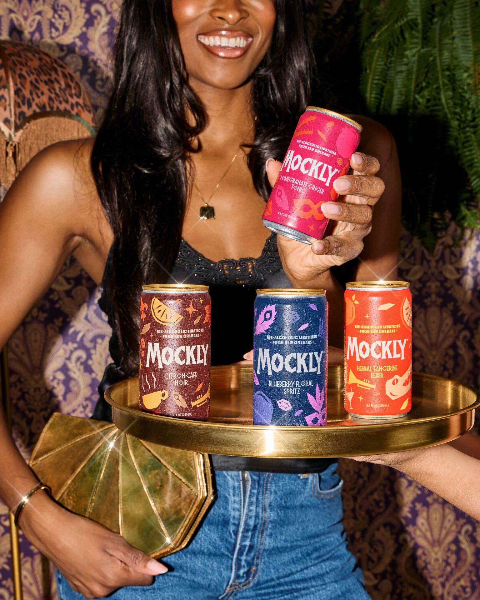 Smiling woman holds a pink Mockly non-alcoholic drink, with three more Mockly cans on a golden tray.