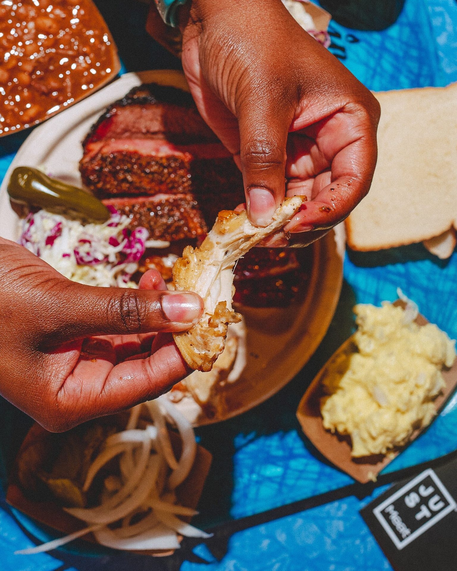 A person's hands pull apart BBQ chicken over a plate of brisket and sides.