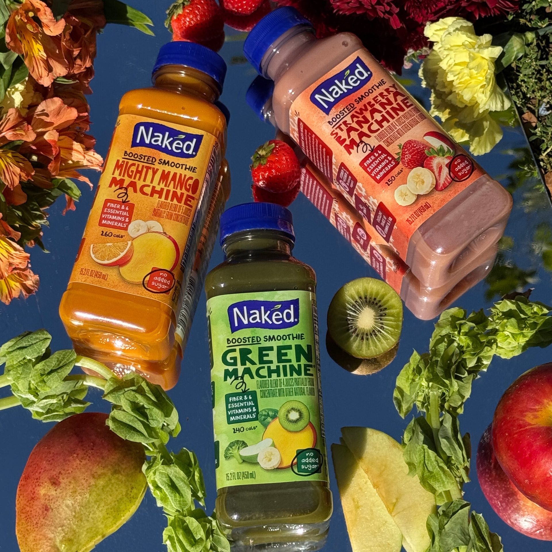 Three Naked smoothies (Mango, Green, Strawberry Banana) with fruit & flowers on a reflective surface.