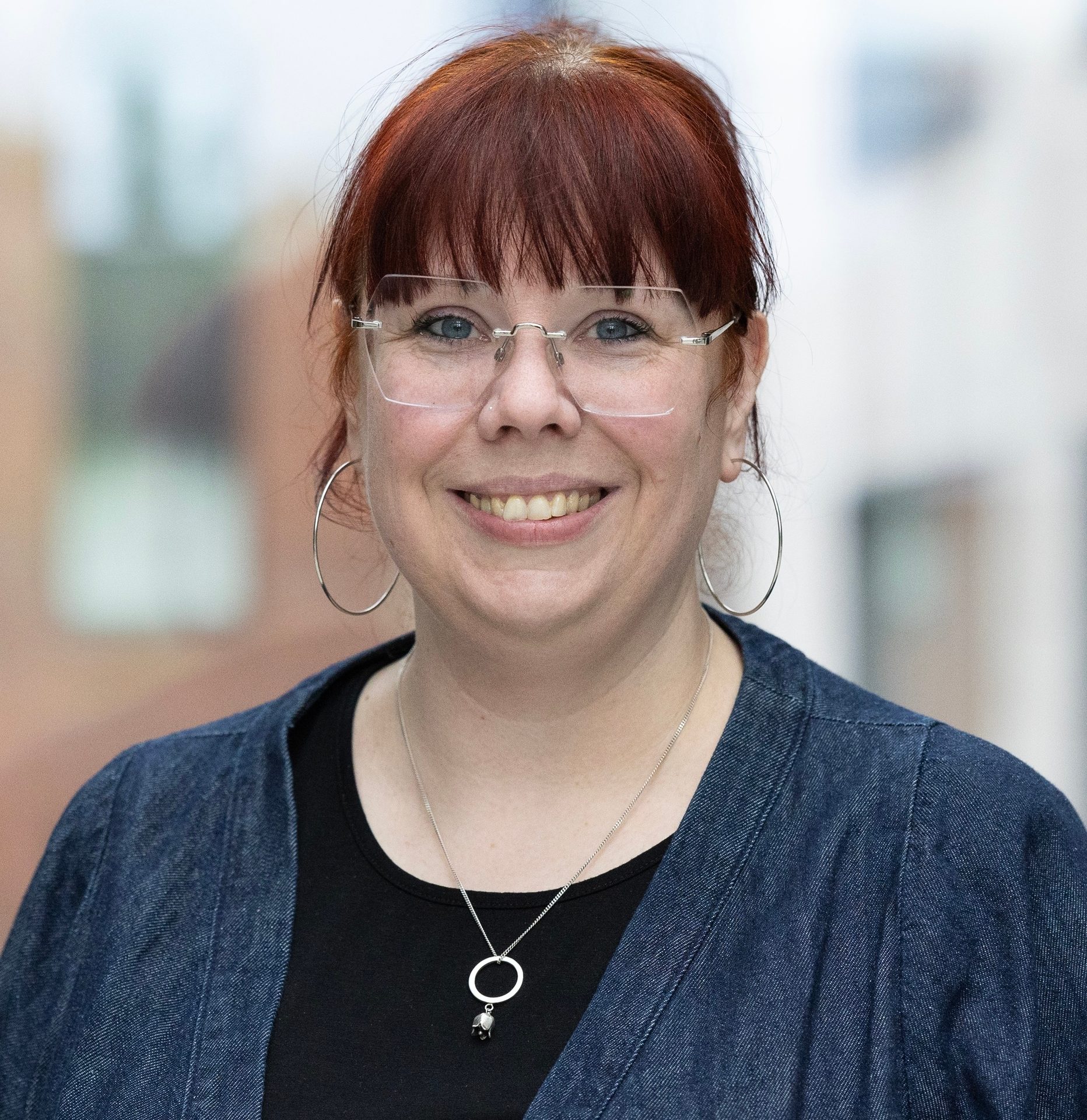 Smiling woman with red hair and glasses, hoop earrings, and a necklace, wearing a black top and blue jacket.