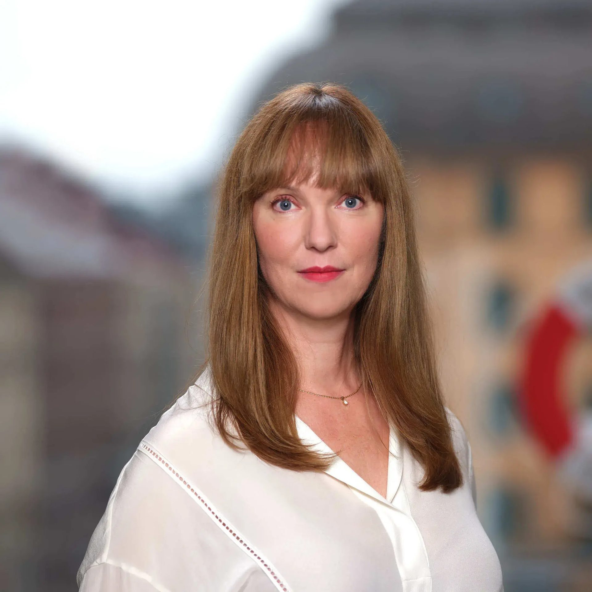 Headshot of a woman with long auburn hair and blue eyes in a white blouse, blurred city background.