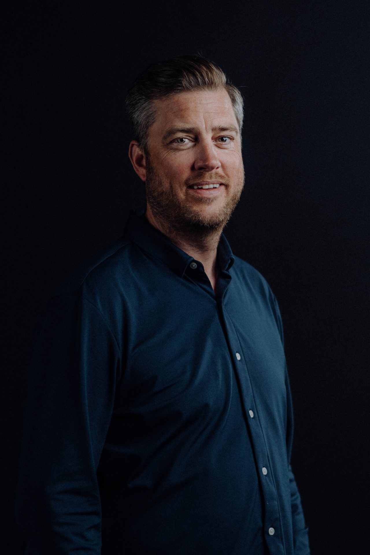 Smiling man with graying hair and beard in a blue shirt against a dark background.