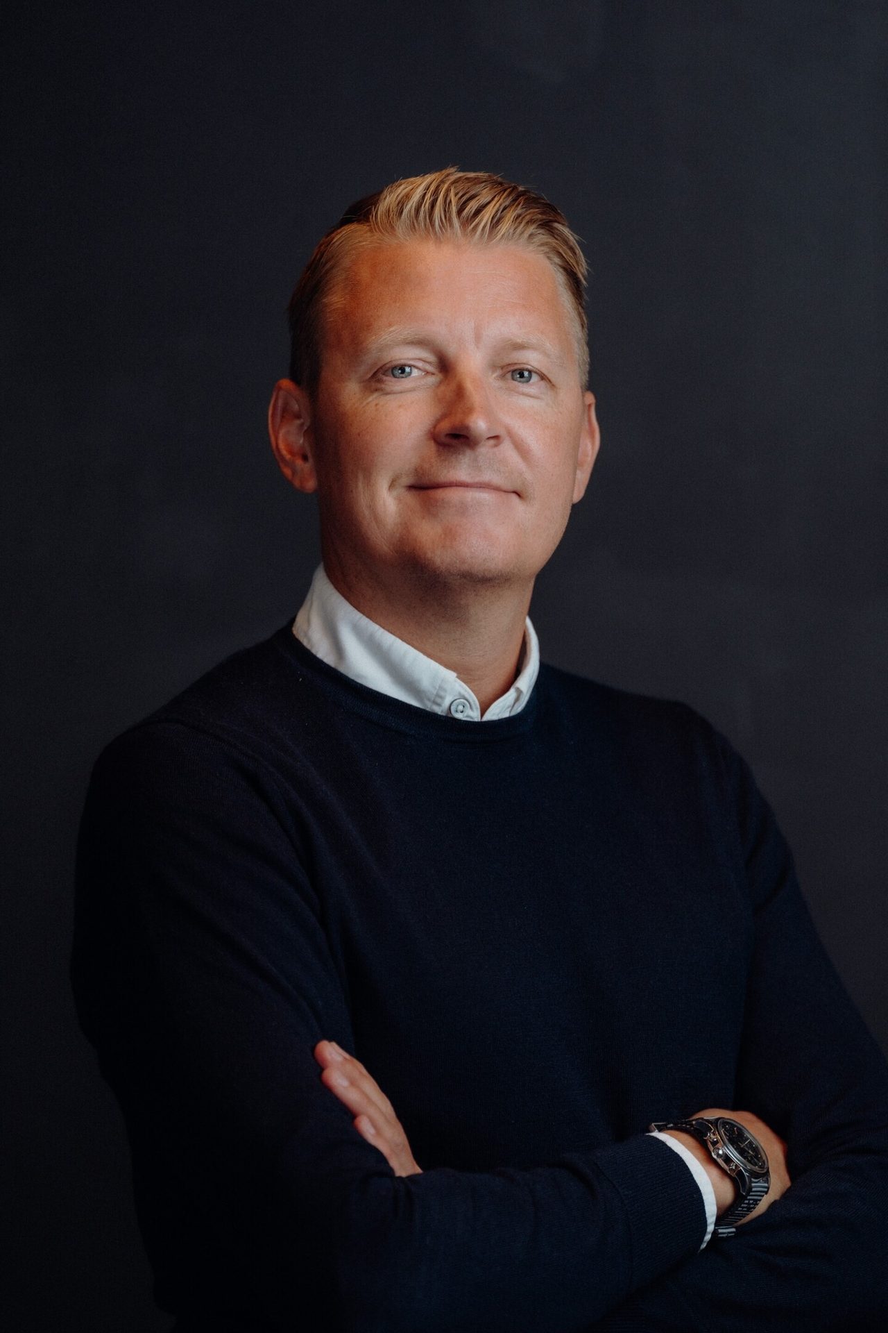 Smiling man, blond hair, blue eyes, dark sweater, white shirt, arms crossed.