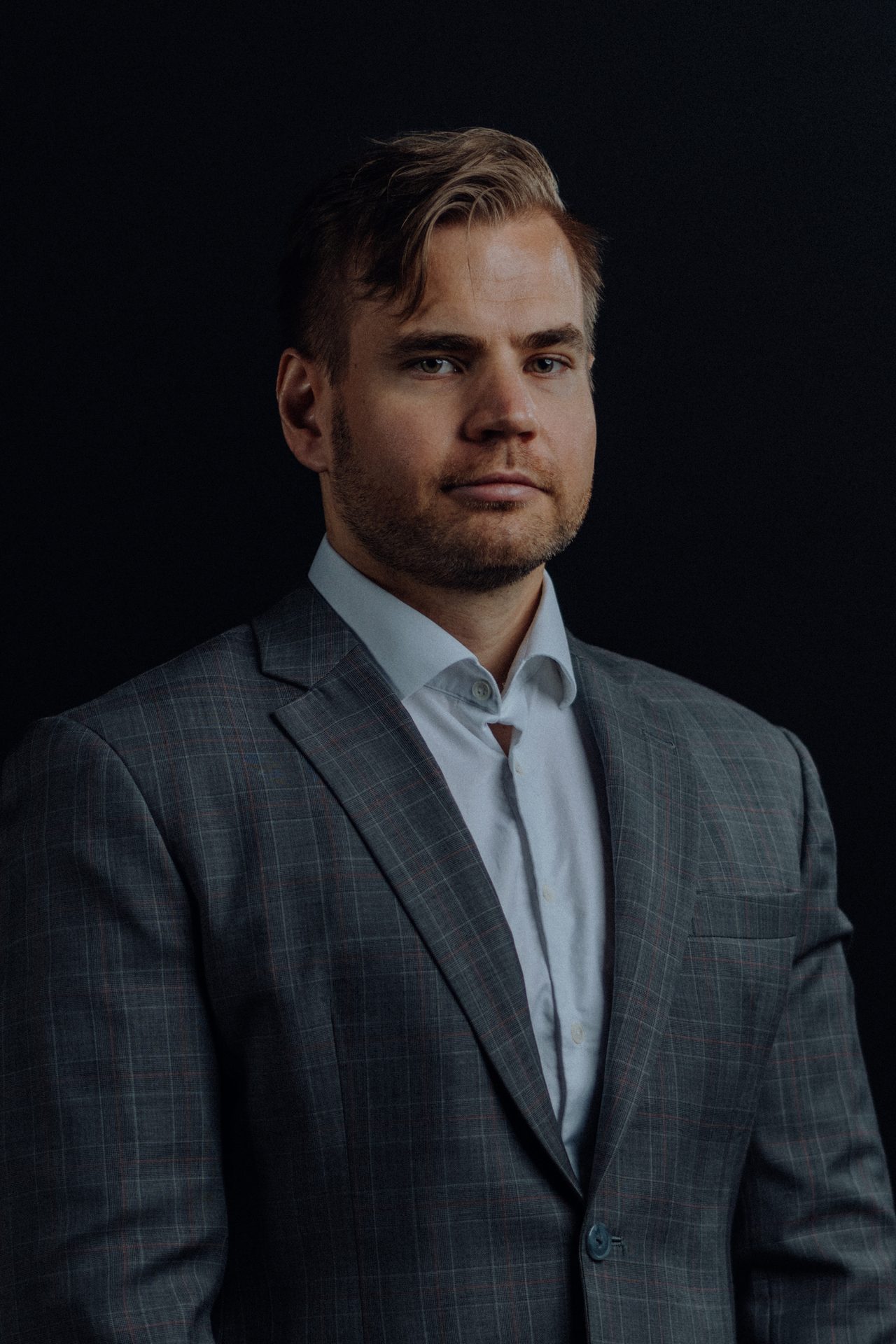 Portrait of a man in a grey plaid suit with a serious expression, dark background.