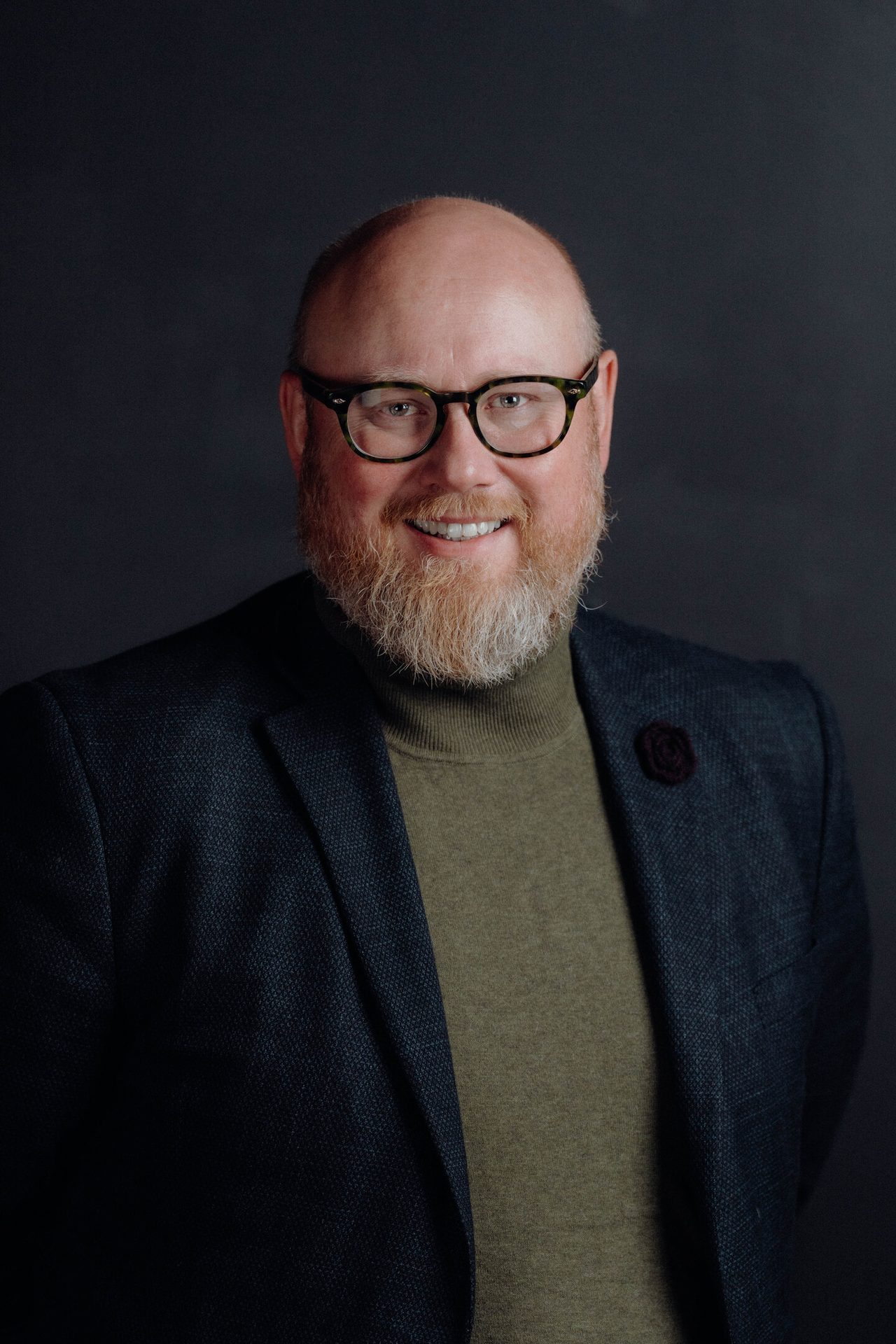 Smiling bald man with beard, glasses, dark jacket, olive turtleneck on a dark background.