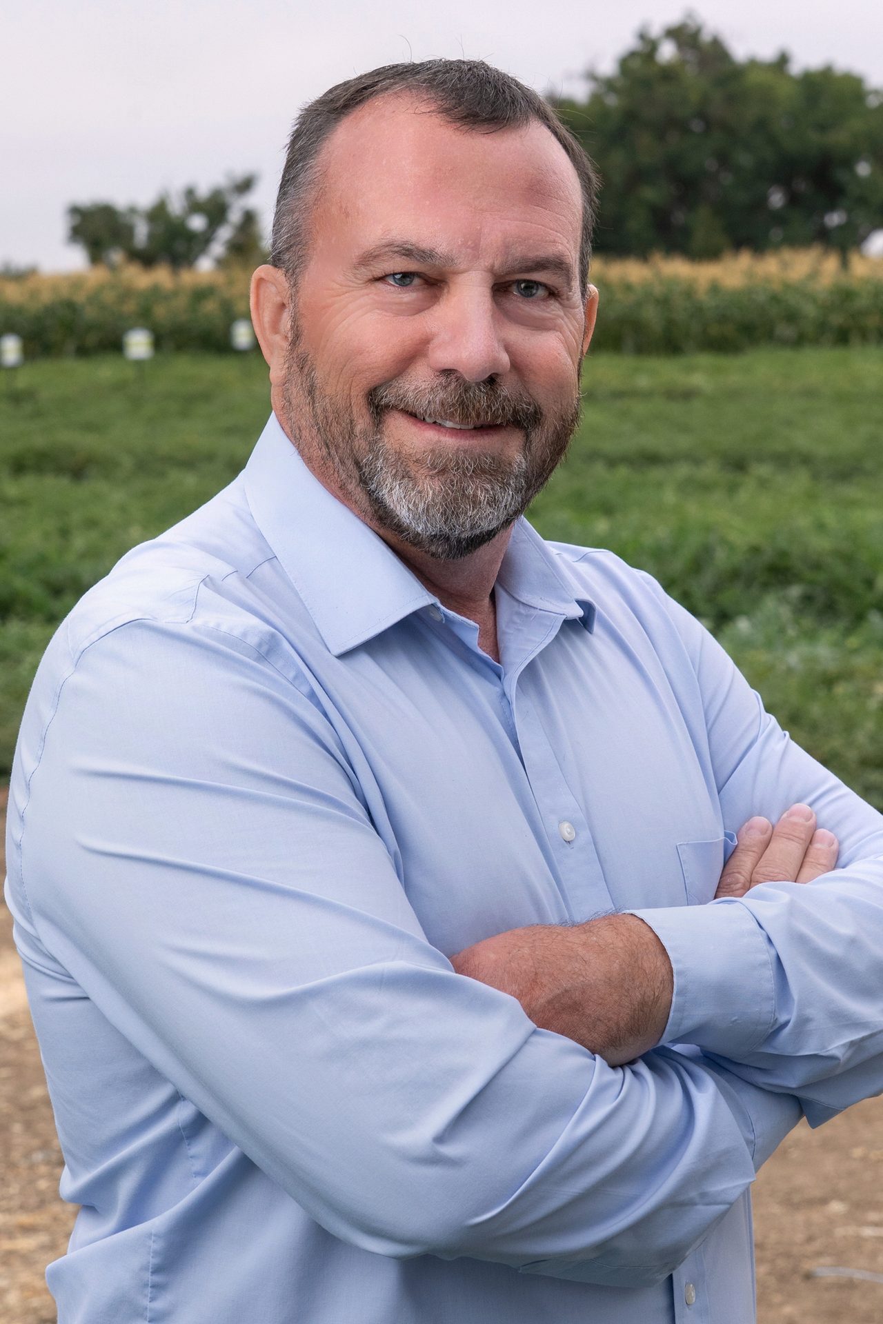 A smiling man with a beard, wearing a light blue shirt with crossed arms, standing in a green field.